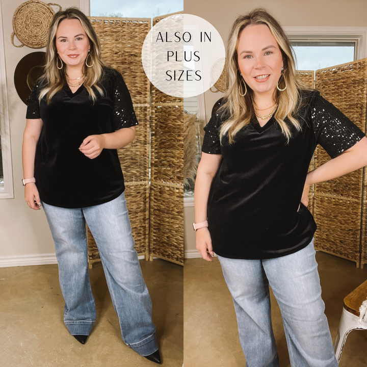 Model is wearing a black velvet short sleeve shirt with black sequins on the sleeve.  Model has long, light wash trouser jeans and black booties. Model has paired this outfit with gold jewelry. Background is brown. 