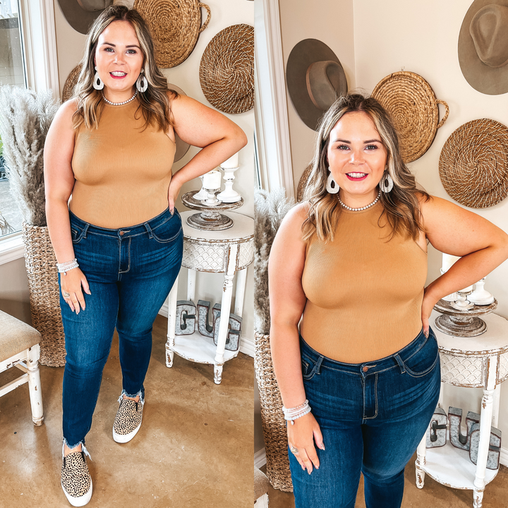 Model is wearing a high neck tank top bodysuit that is caramel brown. Model has it paired with skinny jeans. cheetah print sneakers, a pearl necklace, and ivory earrings.