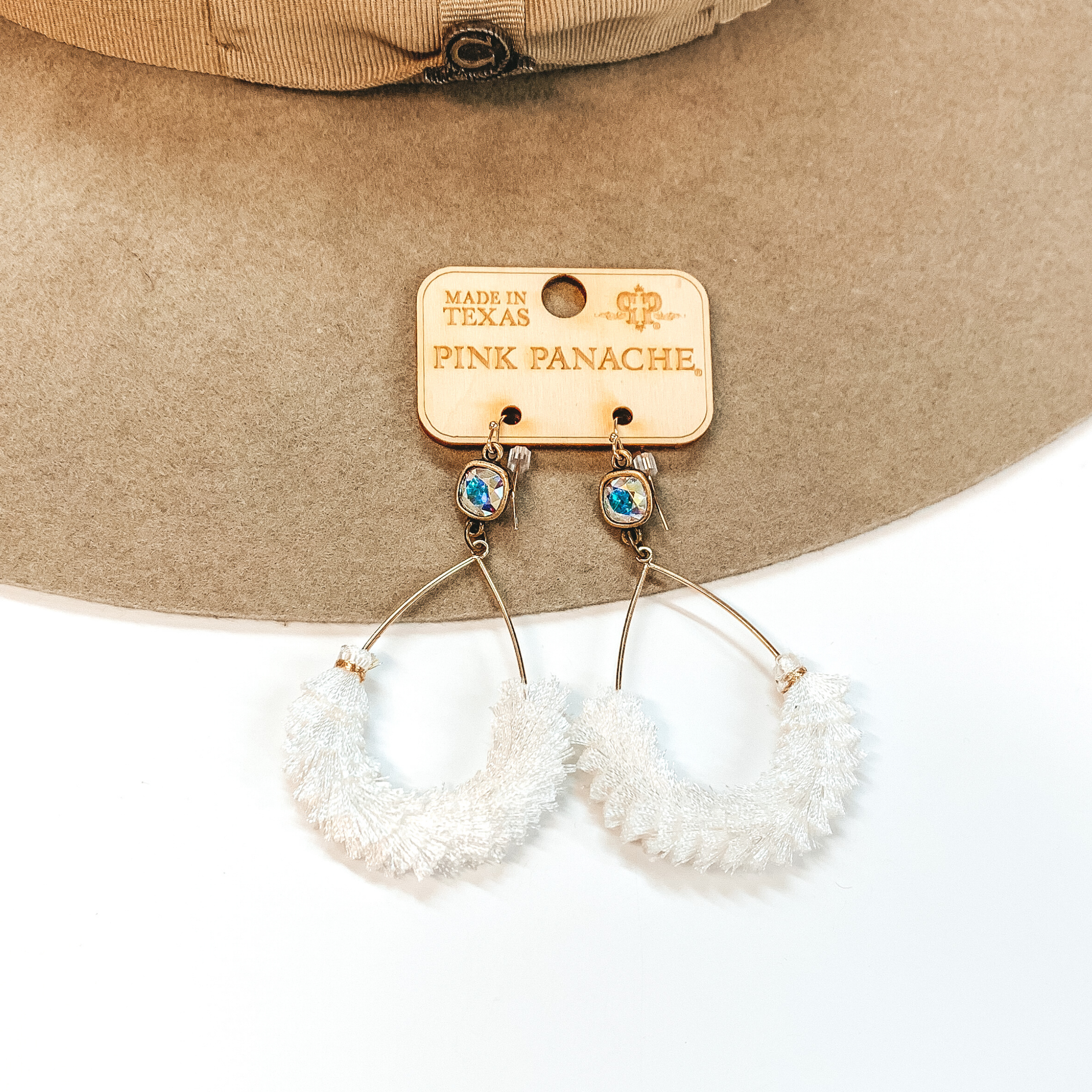 A pair of gold teardrop earrings with an AB cushion cut crystal and white fringe detailing. Pictured on white background with a beige hat.