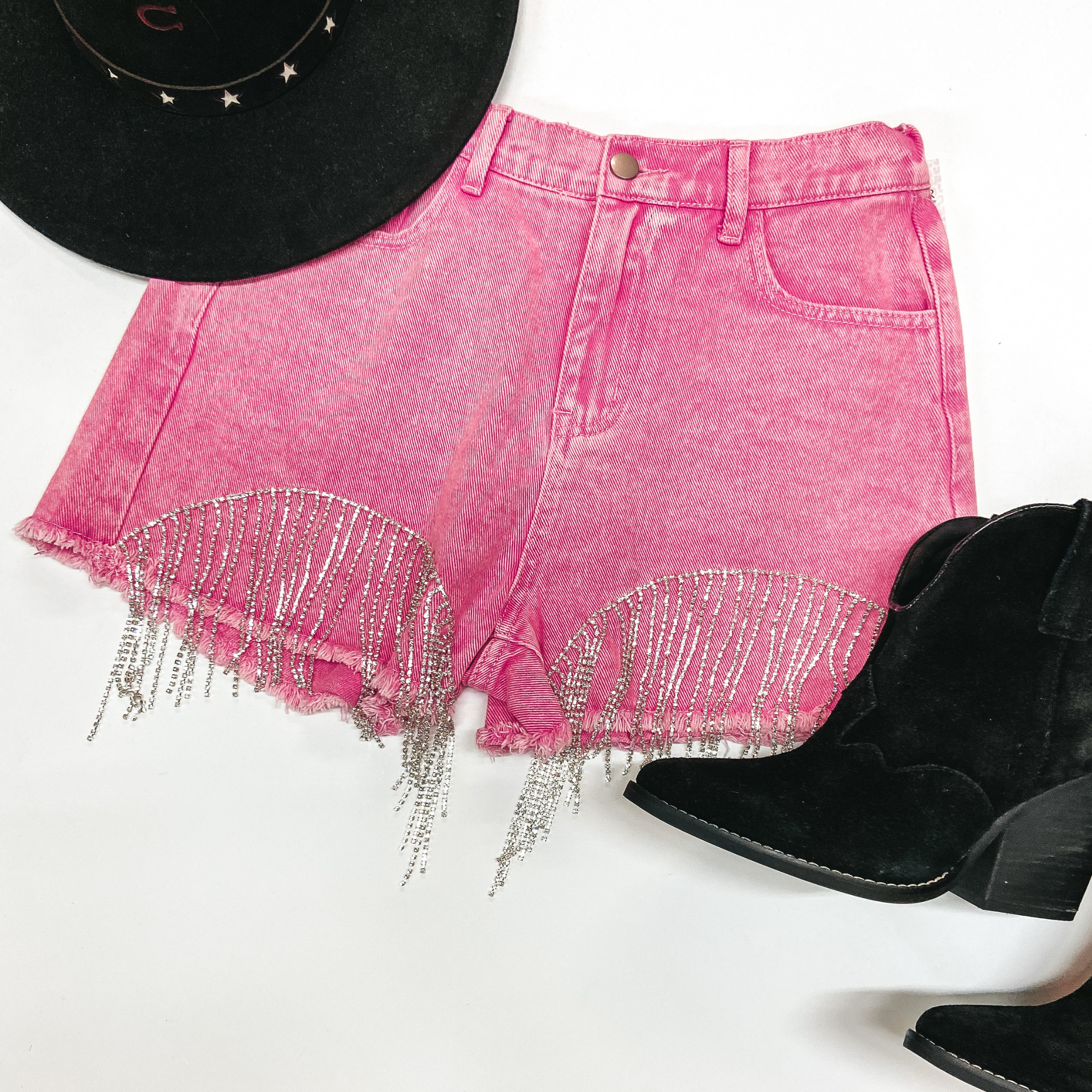 Pink denim shorts with crystal fringe on the bottom on a white background. Paired with a black Charlie 1 Horse hat in the top left and black booties on the bottom right.