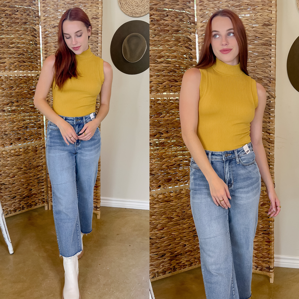Model is wearing a high neck, ribbed sweater, tank top in mustard yellow. The model is wearing light wash jeans and white boots.