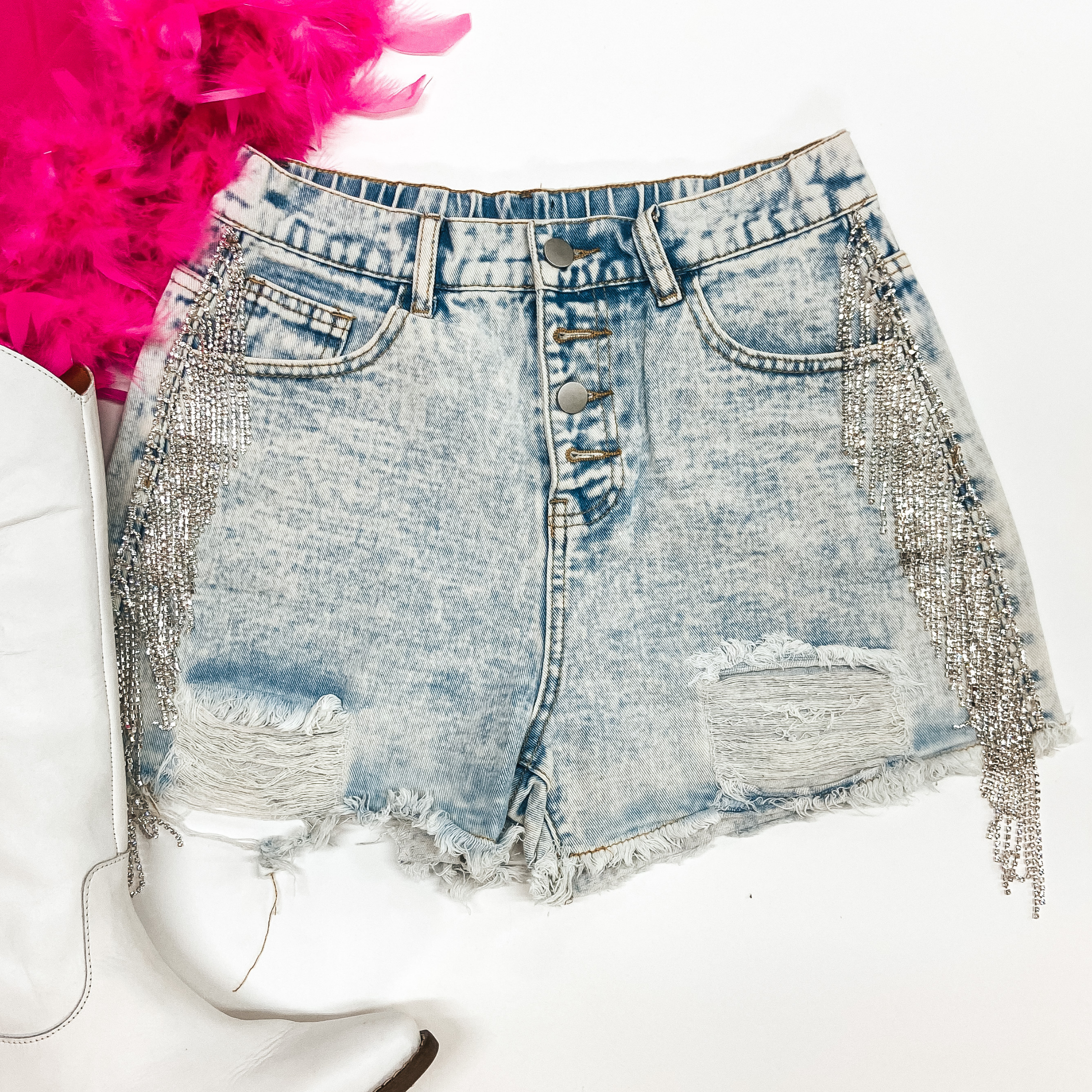 A pair of distressed light wash shorts with crystal fringe on the hips. These button fly shorts are pictured on a white background with white boots and  a pink feather top.