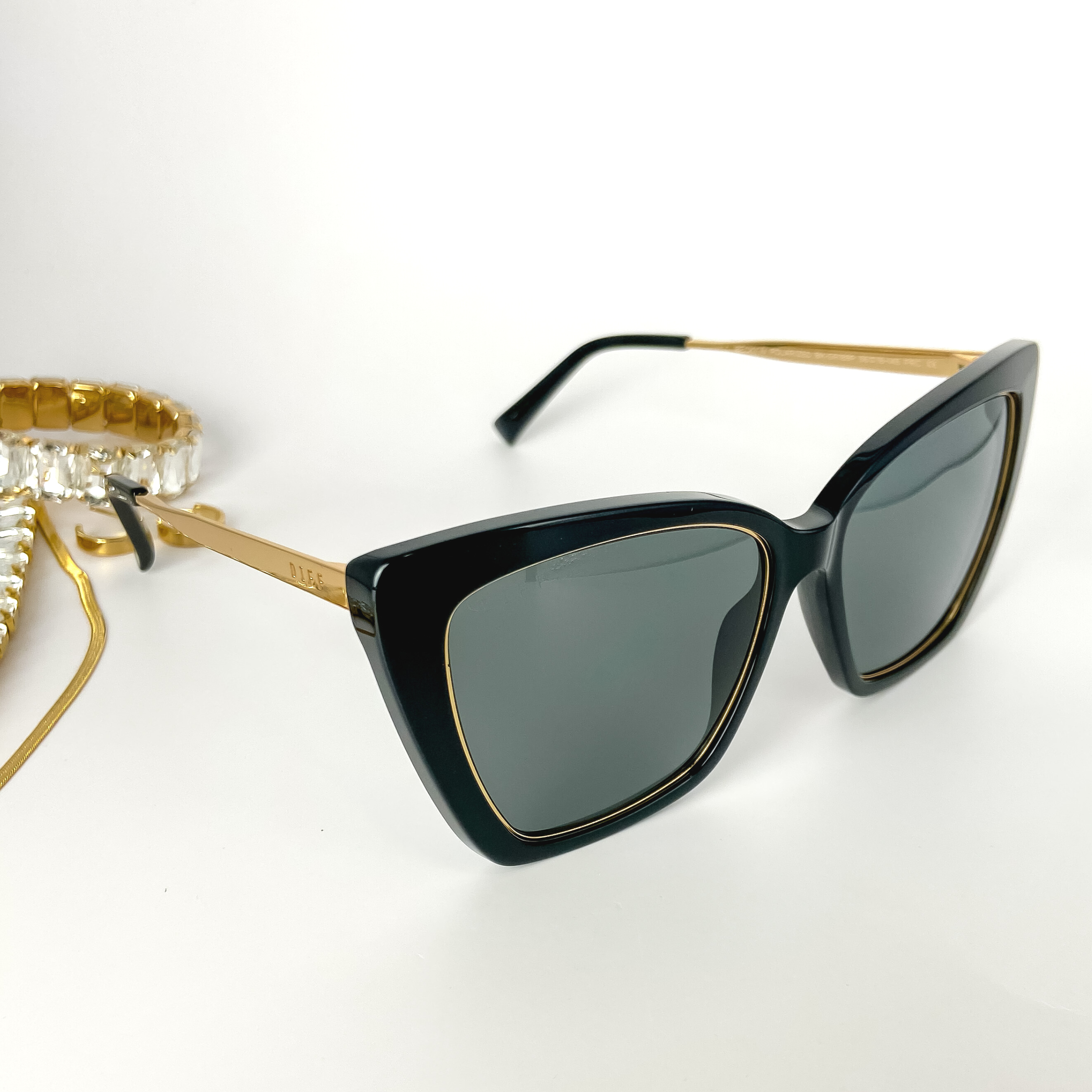 A pair of gold-tone and black cat-eye sunglasses pictured open on a white background with gold jewelry.