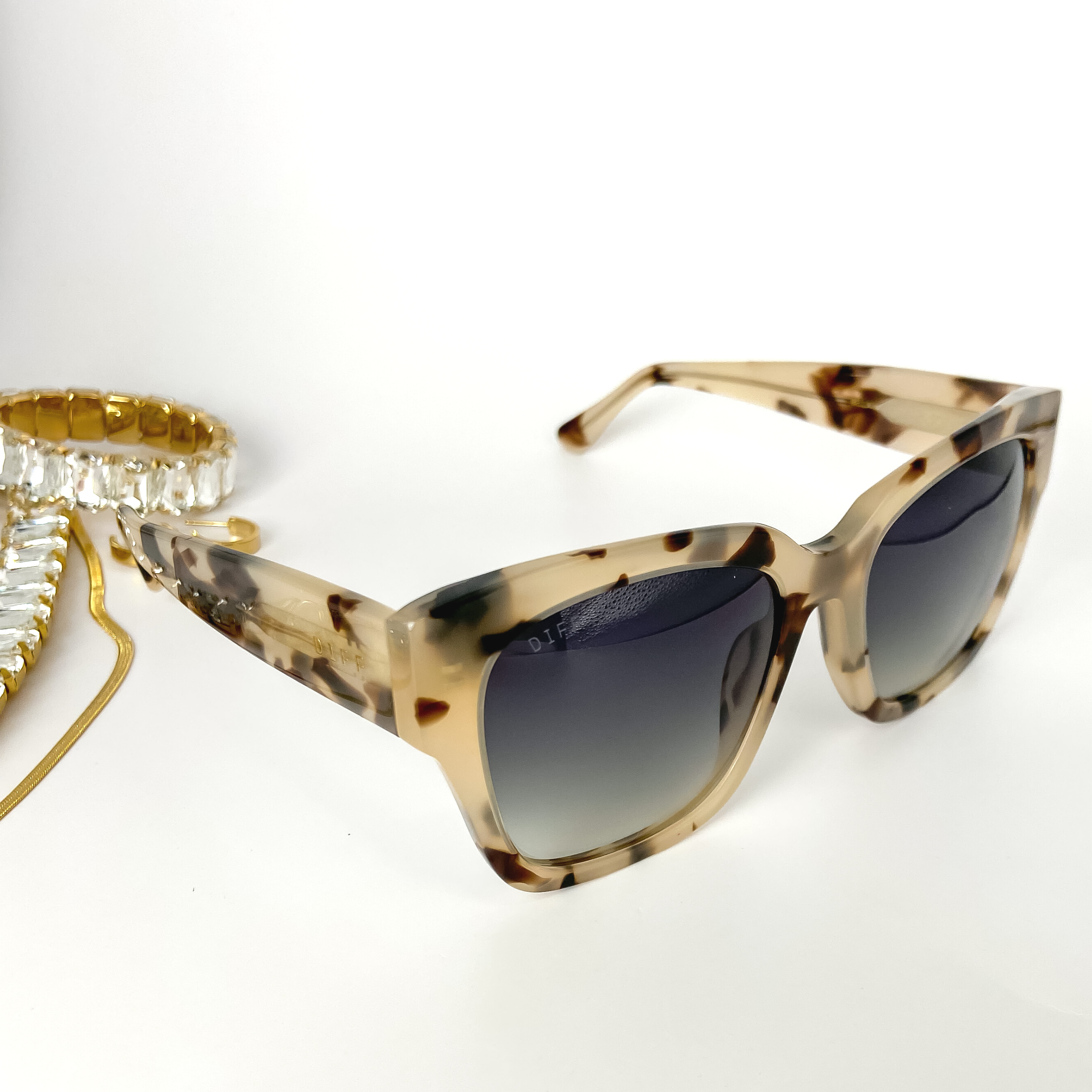 A pair of cream tortoise-print square sunglasses with grey fade lenses pictured open on a white background with gold jewelry.