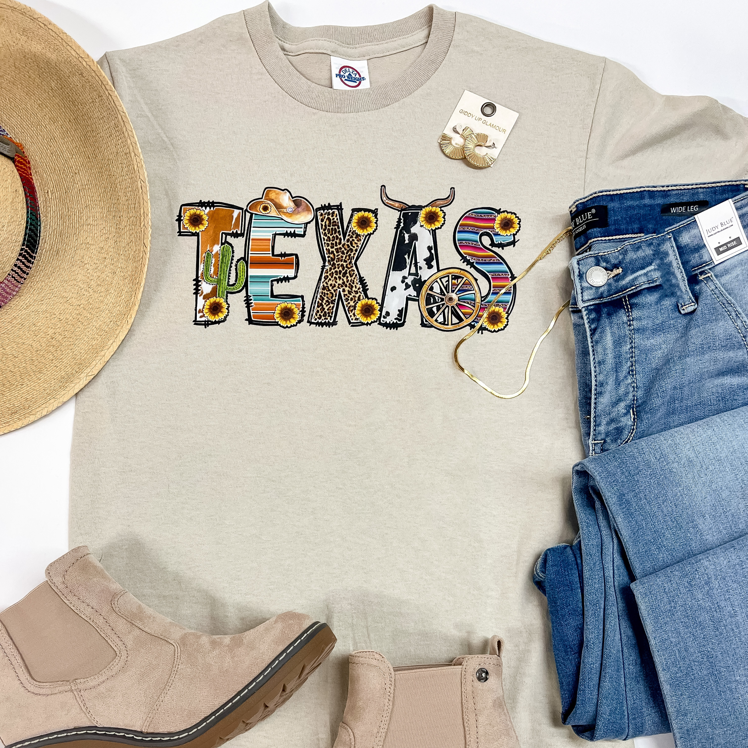 A beige tee with "Texas" written in block letters in the middle. The prints inside "Texas" are, but not limited to: Cowhide, serape, leopard. On the left of the tee is a straw Charlie 1 Horse hat and brown booties. To the right is a pair of blue jeans, gold hoop earrings, and a gold necklace