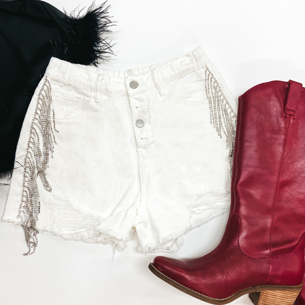 A pair of white denim shorts with a button fly and crystal fringe on the hips. These shorts are pictured on a white background with red boots and black feather top.