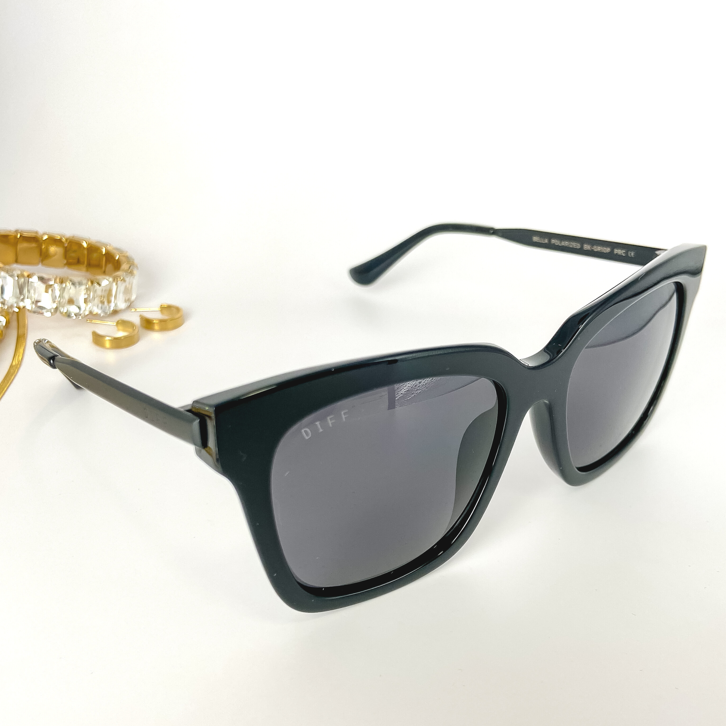 A pair of black square sunglasses with black lenses pictured open on a white background with gold jewelry.