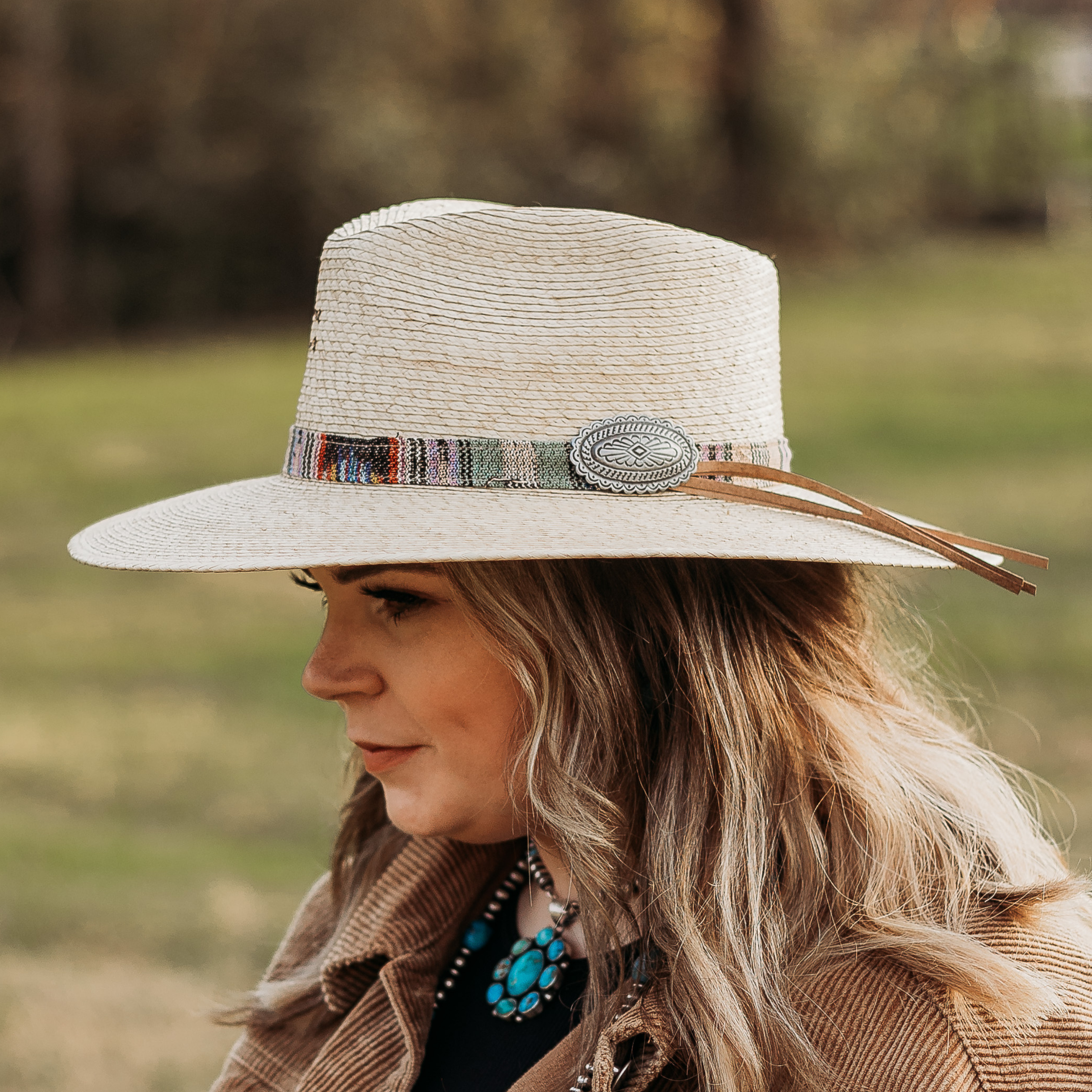 Off White Straw Hat. Multi-Colored Band with Silver Concho Pin and Brown Leather Detailing. Model has it paired with a brown jacket and turquoise jewelry. Pictured on wooded background.