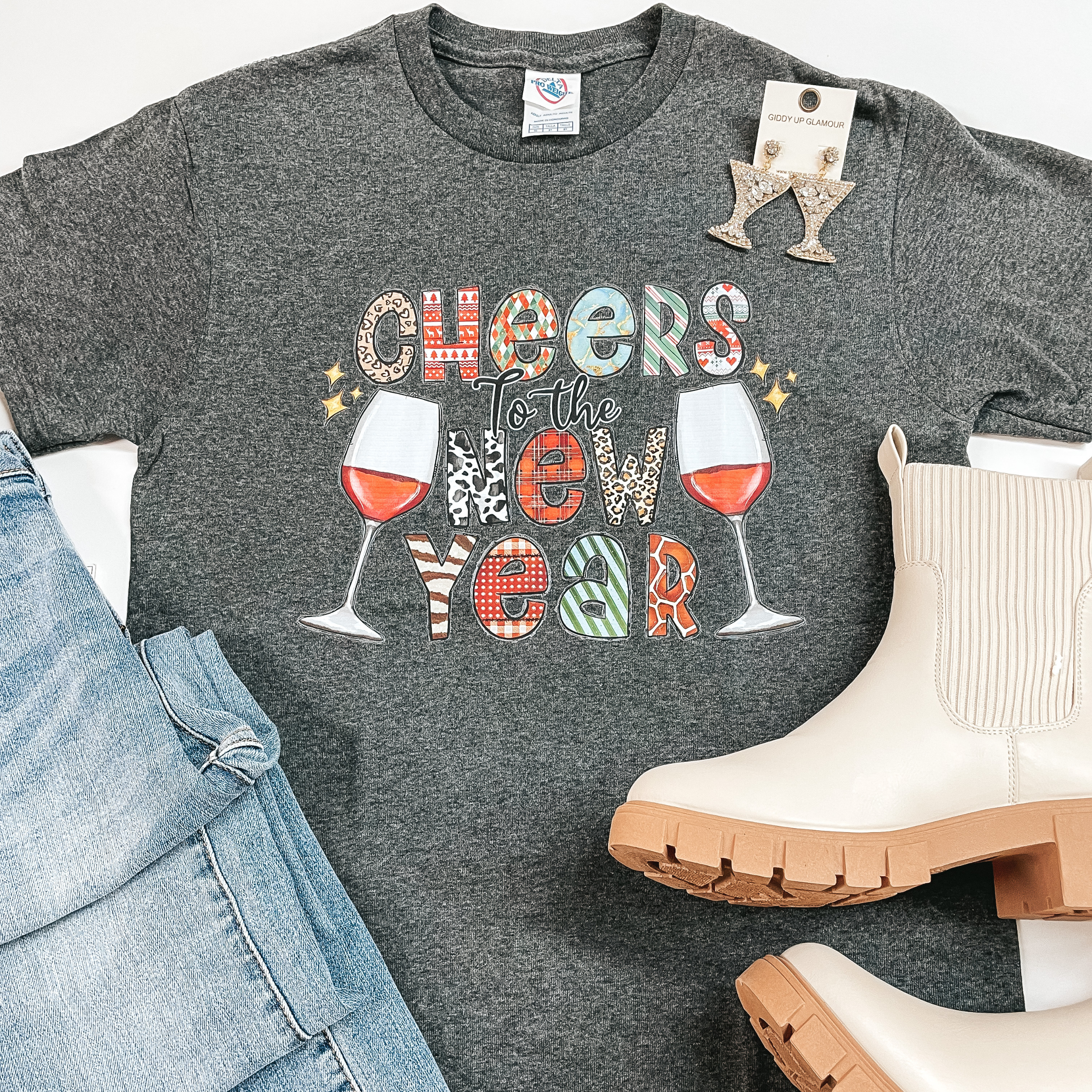 A gray tee with "Cheers To The New Year" in the middle with various prints within the letters. On each side of the saying there are wine glasses half full. On the bottom left there is a light-wash pair of jeans and to the bottom right there are cream booties. A pair of earring is laid on the top right of the tees shoulder. 