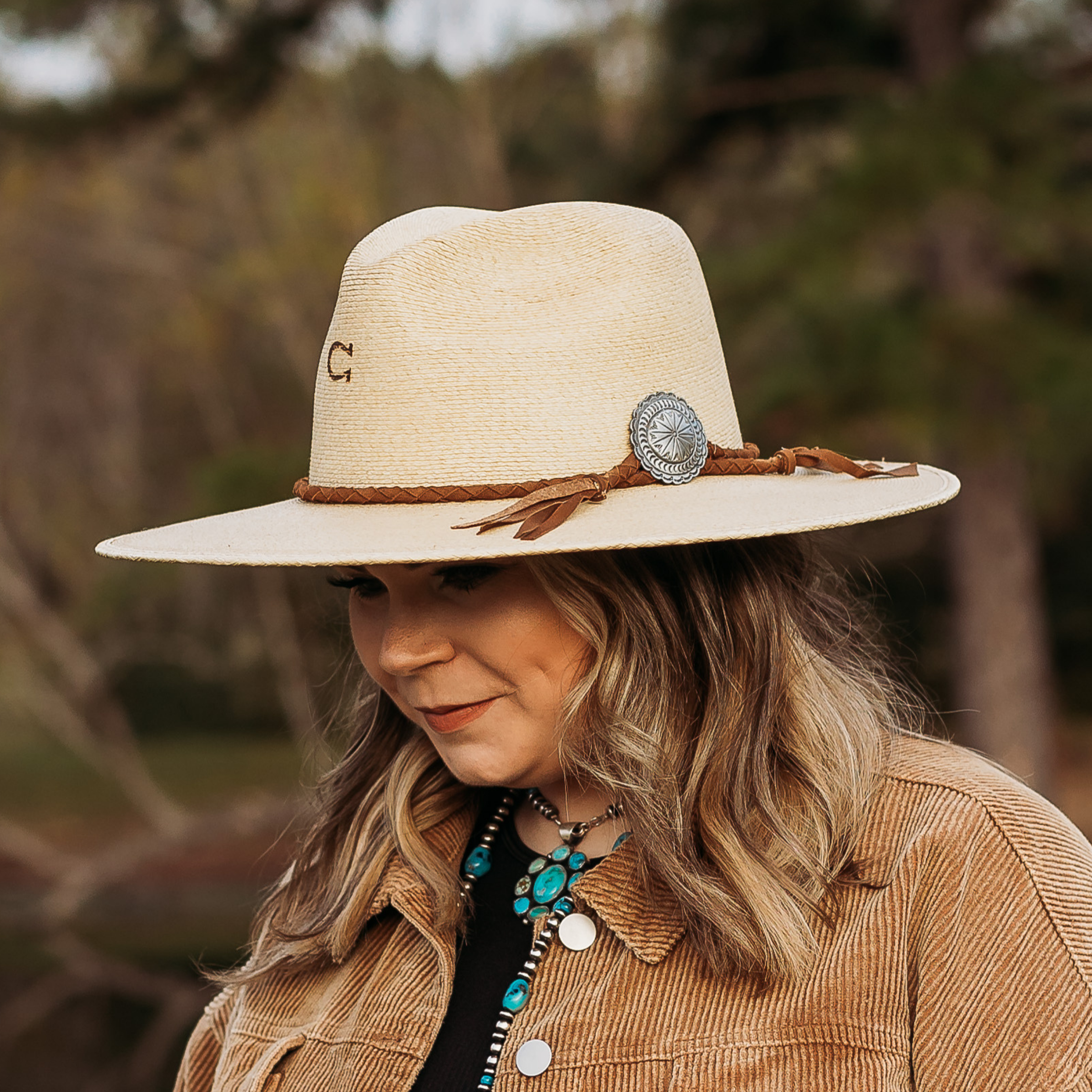 Off White Straw Hat with Braided Tan Hat Band, and a Silver Concho.. Model has it paired with a brown jacket and turquoise jewelry. Pictured on wooded background.
