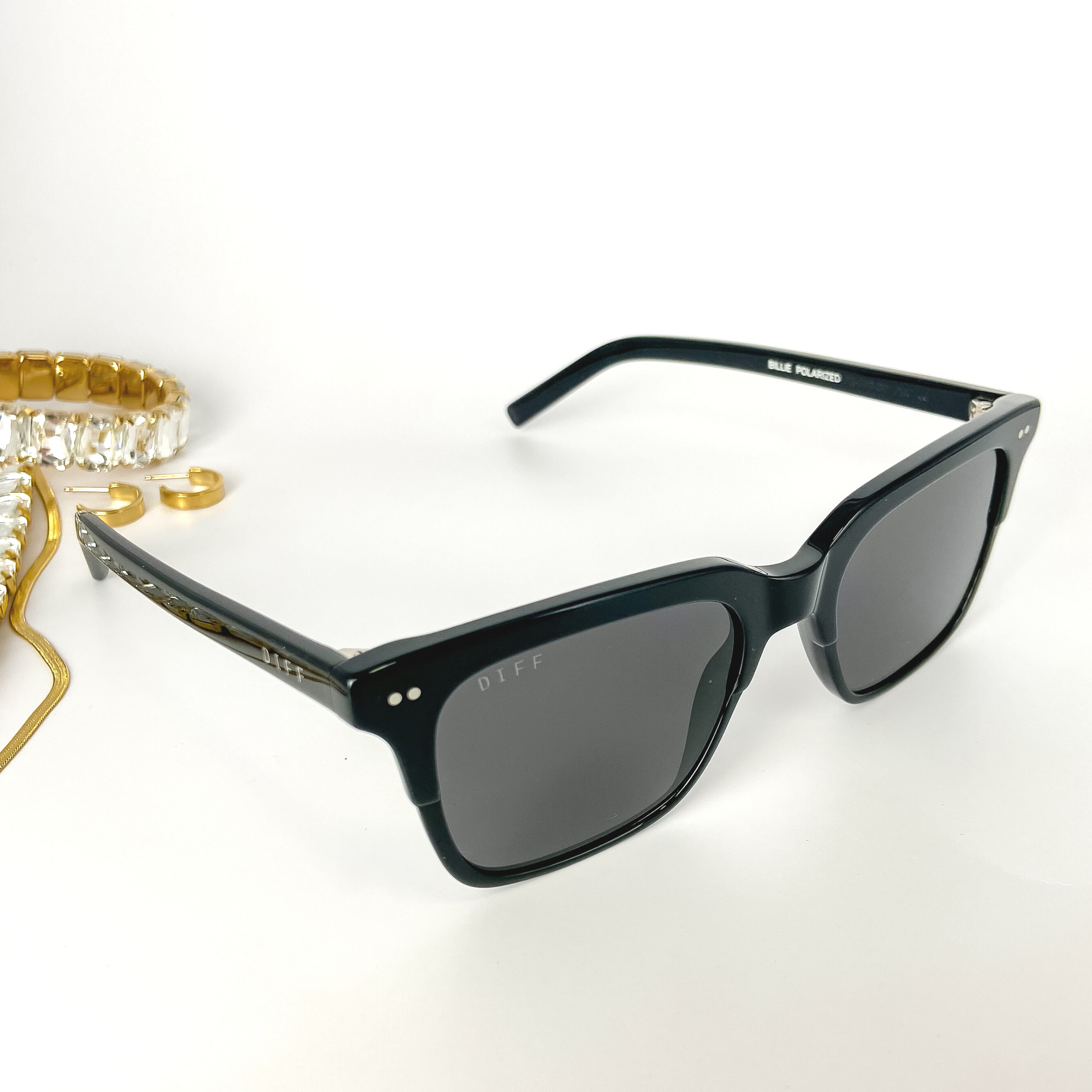 A pair of black sunglasses with black lenses. These sunglasses are pictured on a white background with gold jewelry.