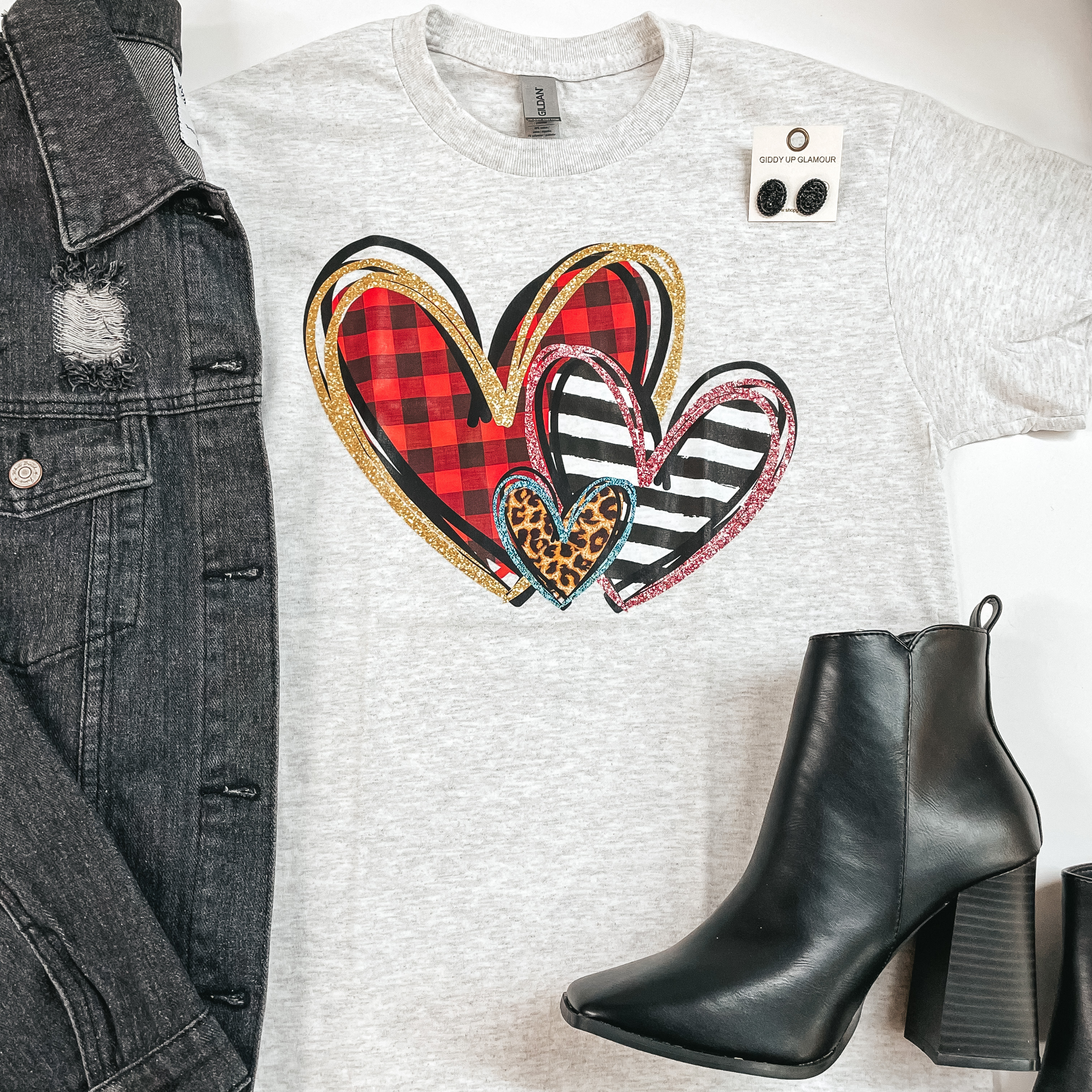 A gray tee with three various sized hearts in the middle with designs including: red buffalo plaid, black and white stripes, and cheetah. Pictured on the left is a black denim jacket and to the right is black booties. 