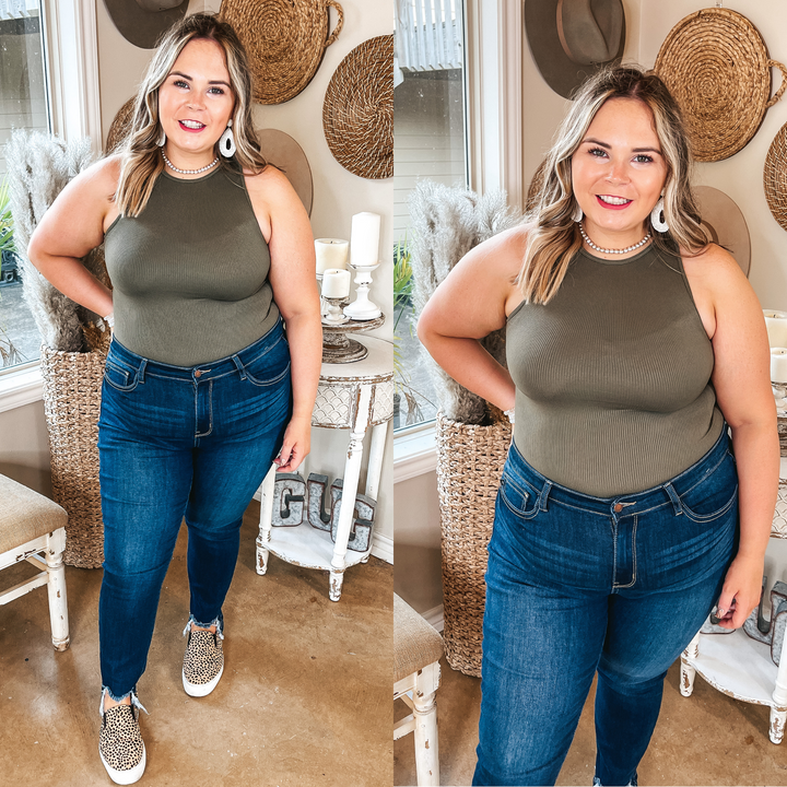 Model is wearing a high neck tank top bodysuit that is olive green. Model has it paired with skinny jeans, cheetah print sneakers, a pearl necklace, and ivory earrings.