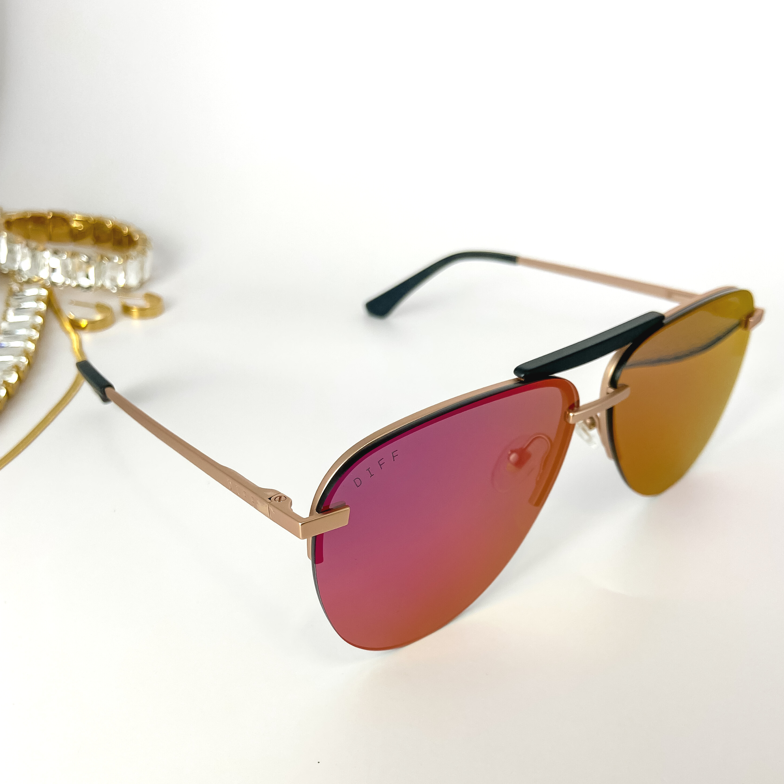A pair of pink lens aviator sunglasses with gold-tone frames. These sunglasses are pictured on a white background with gold jewelry.
