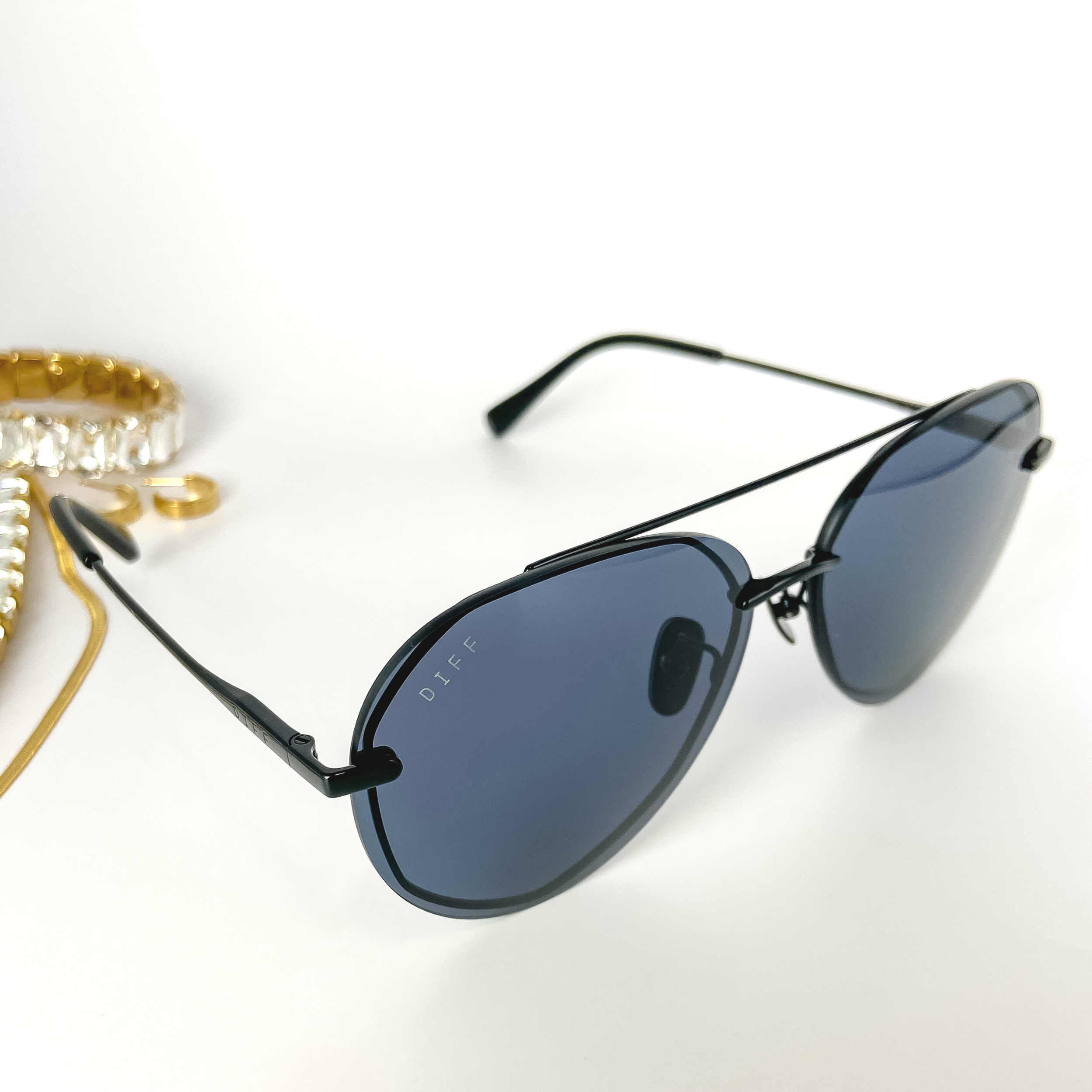 A pair of black aviator sunglasses with grey lenses. These sunglasses are pictured on a white background with gold jewelry.