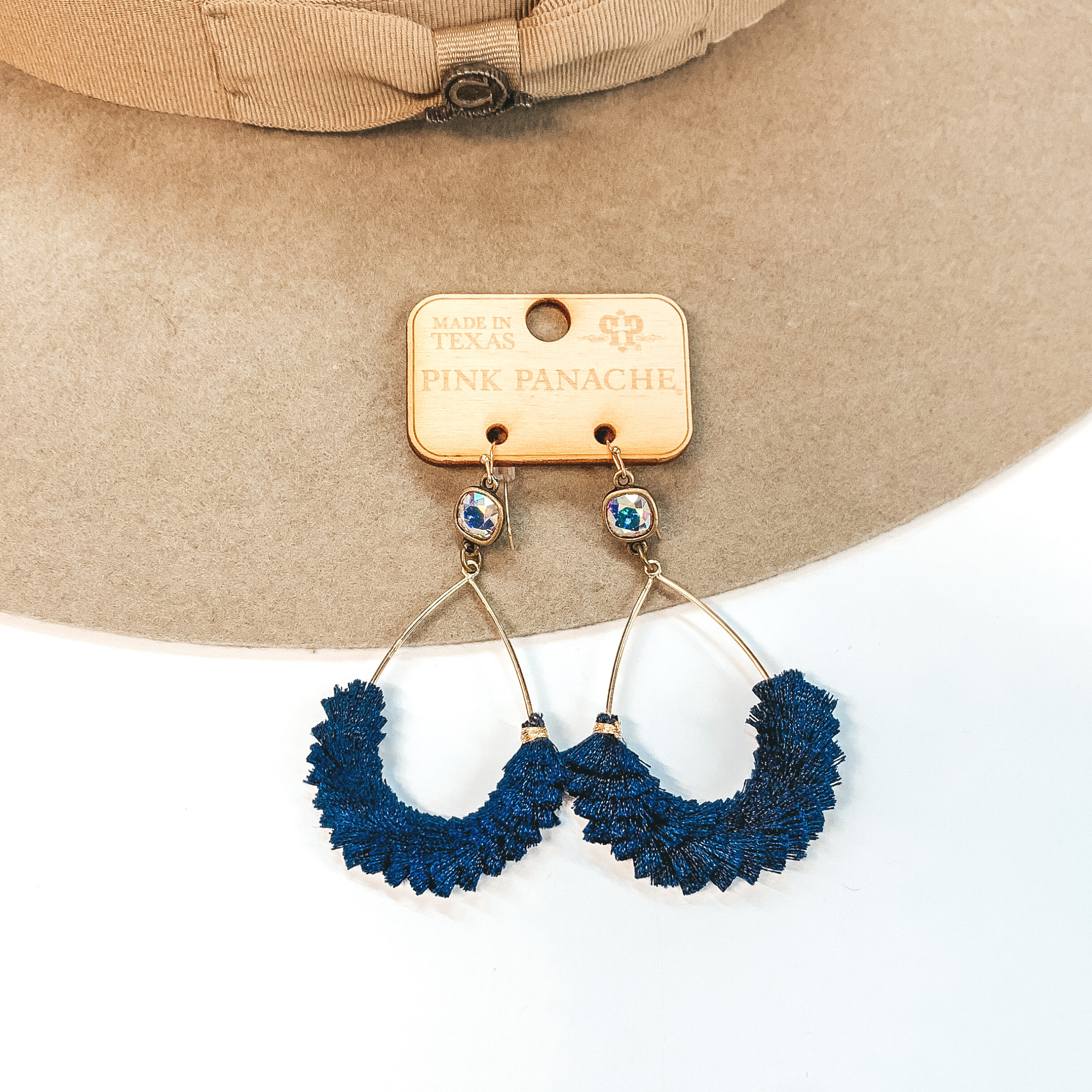 A pair of gold teardrop earrings with an AB cushion cut crystal and navy blue fringe detailing. Pictured on white background with a beige hat.