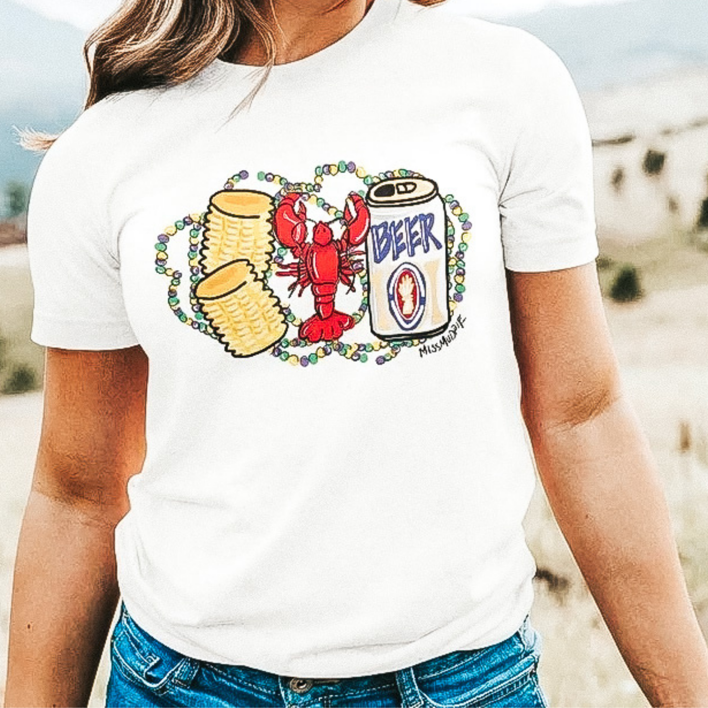 A cream colored crew neck tee shirt. The tee shirt has a graphic of corn, a crawfish, and a beer can. The graphic has mardi gras beads around it.