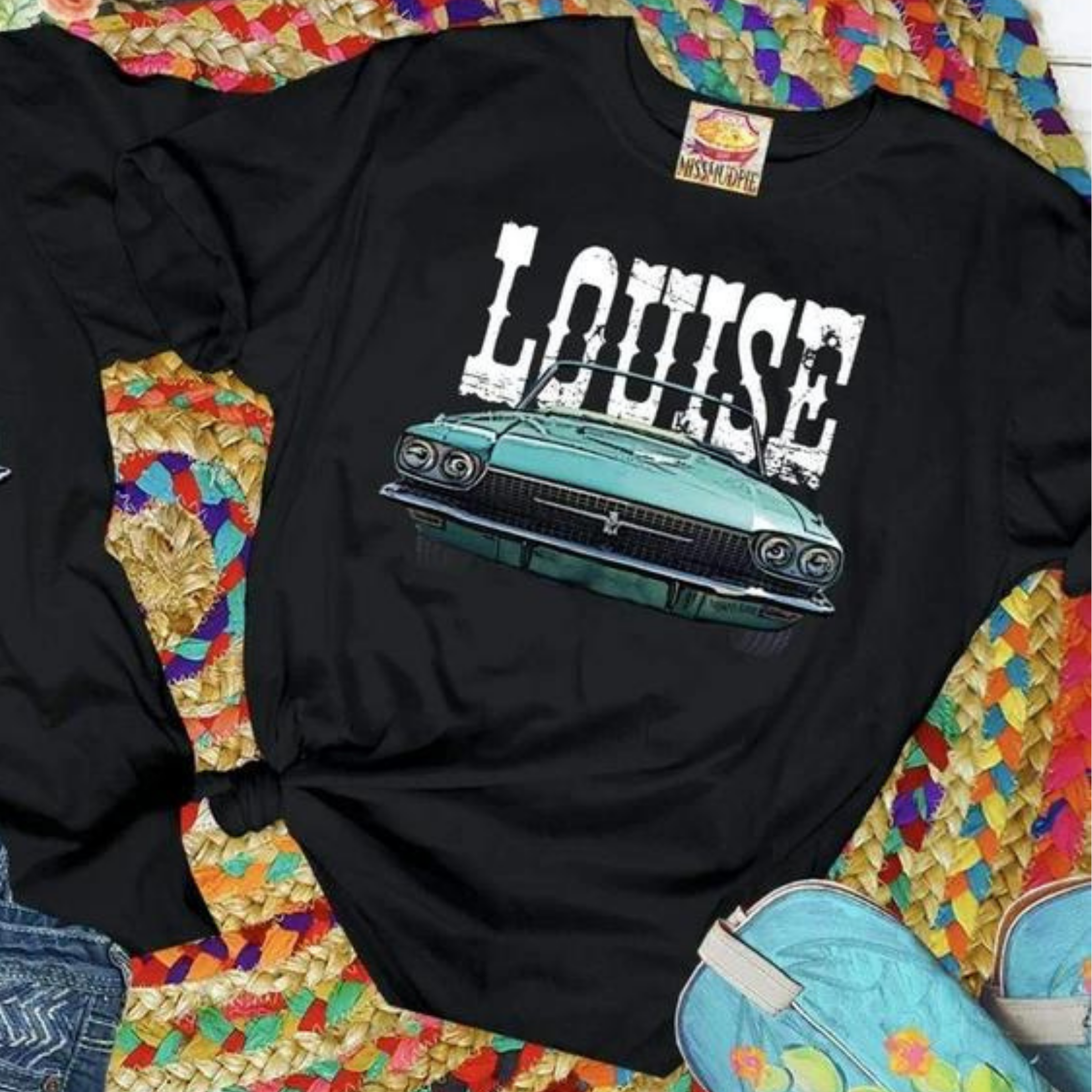 A black tee shirt folded into a rectangle exposing the vintage car graphic that says "Louise" above. Pictured with a turquoise necklace on a serape rug.