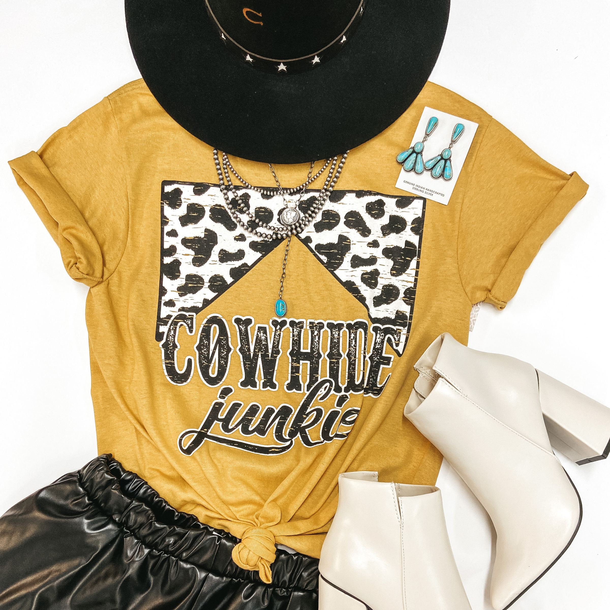 A mustard yellow graphic tee with cuffed sleeves and a knotted front. The graphic has a cowhide print design that says "Cowhide Junkie" underneath. Tee shirt is pictured on white background with ivory booties, black faux leather shorts, a black hat, and sterling silver jewelry.
