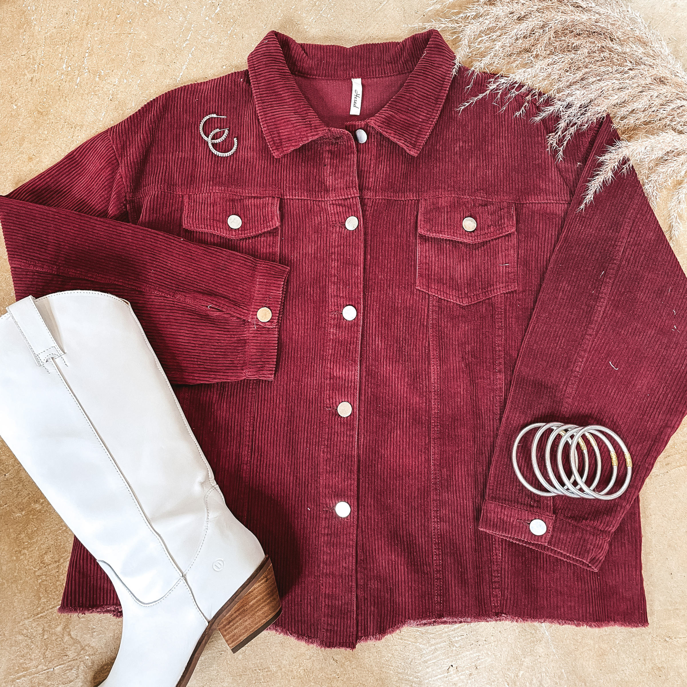 A maroon corduroy shacket pictured on a concrete background with white boots and silver jewelry.