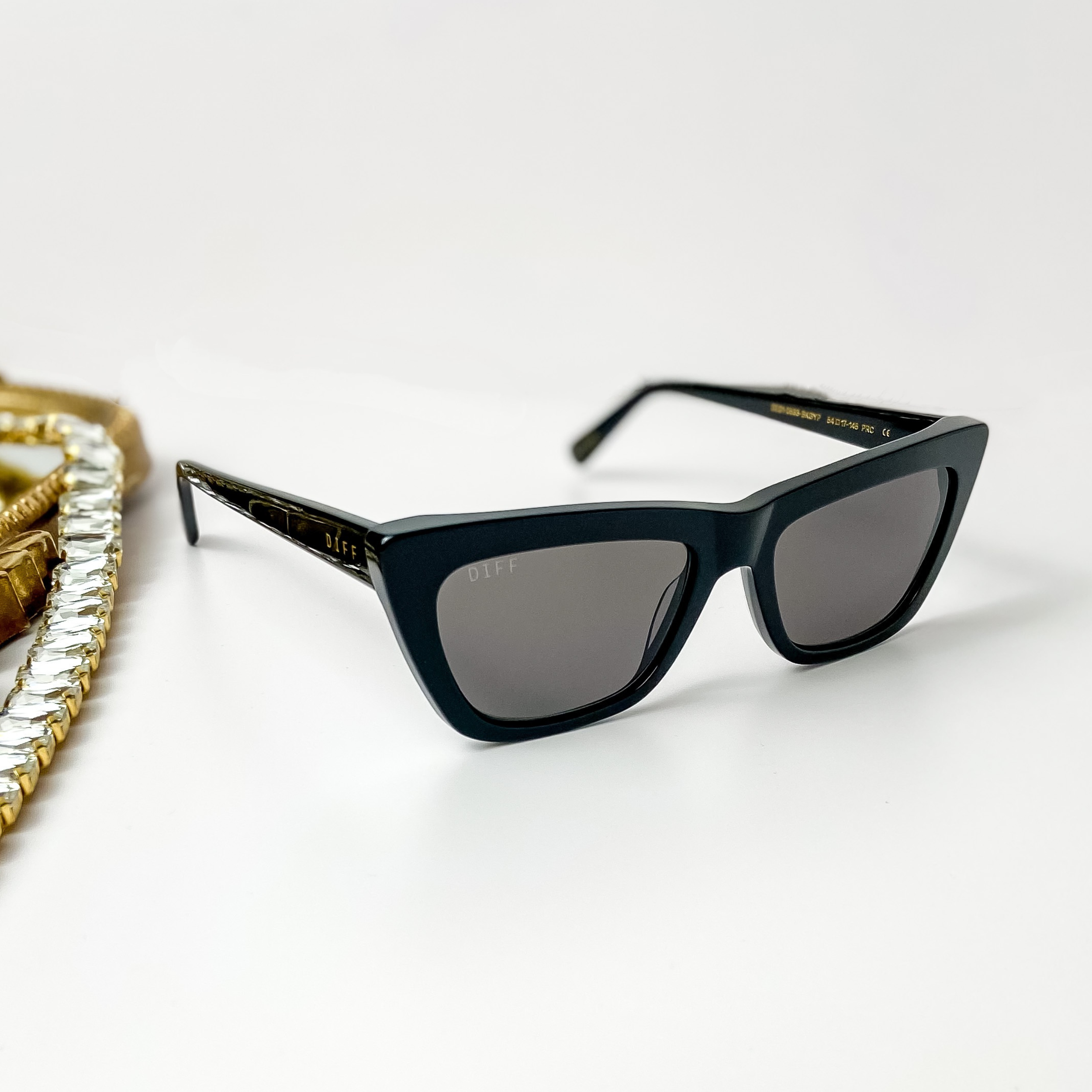 A pair of black cat-eye sunglasses pictured on a white background with gold jewelry.