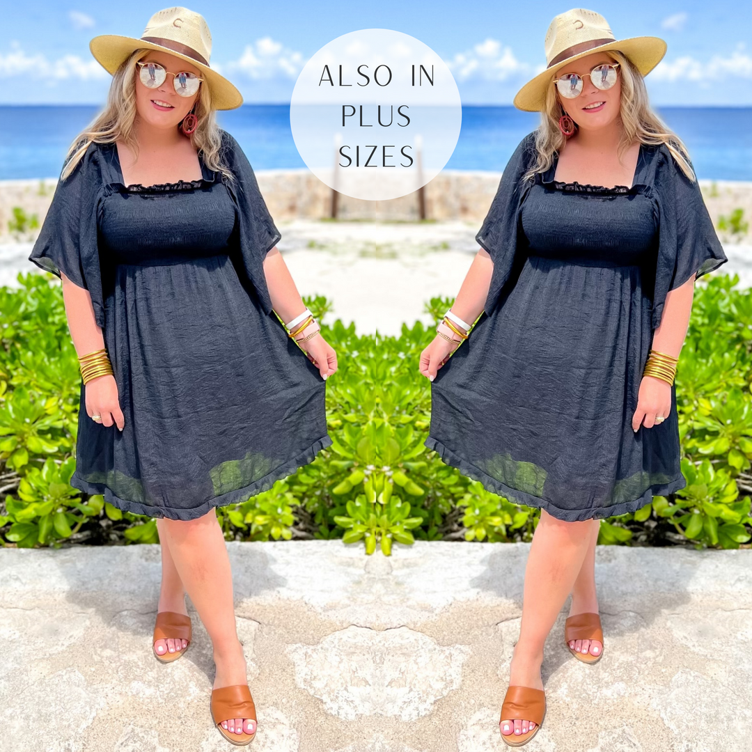 Model is wearing a black dress with ruffle sleeves and a smocked bodice. Model has it paired with a straw hat, tan sandals, and gold jewelry.