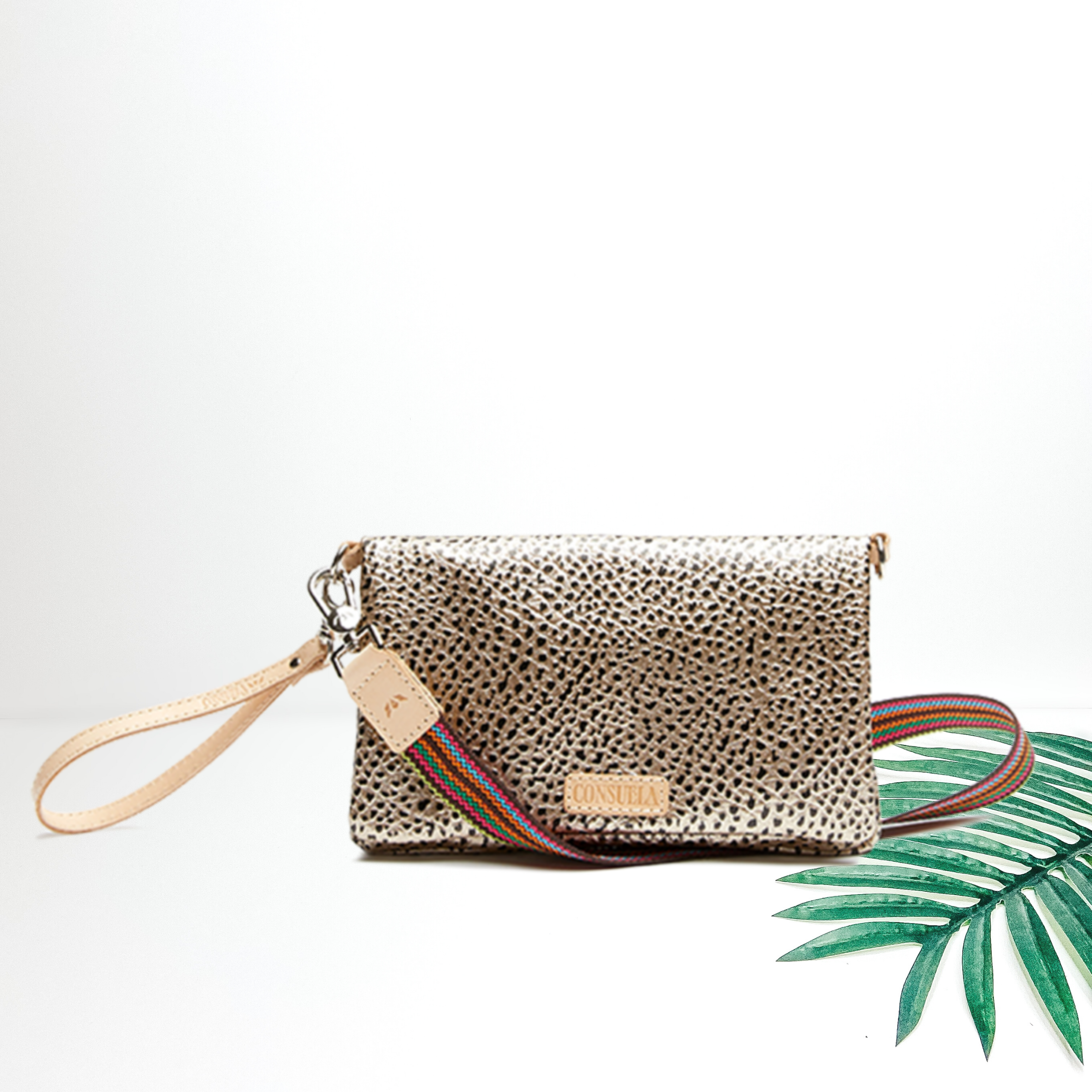 A gold metallic crossbody bag with a leather wristlet strap. Pictured on white background with a palm leaf.