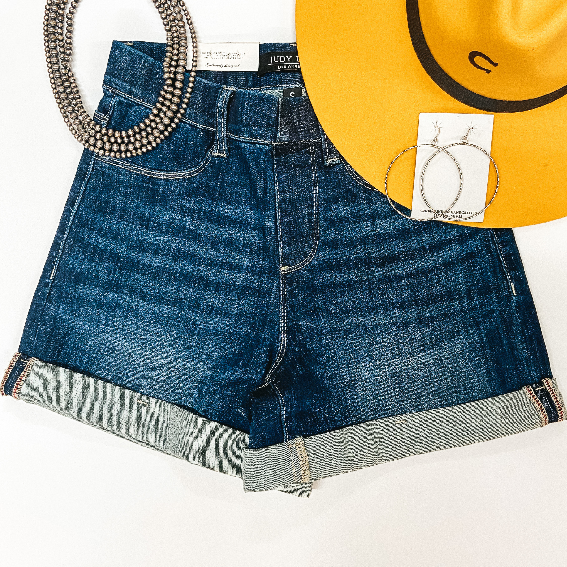 A pair of dark wash elastic waist shorts that have a cuffed hem. Pictured on white background with sterling silver jewelry and a yellow hat.