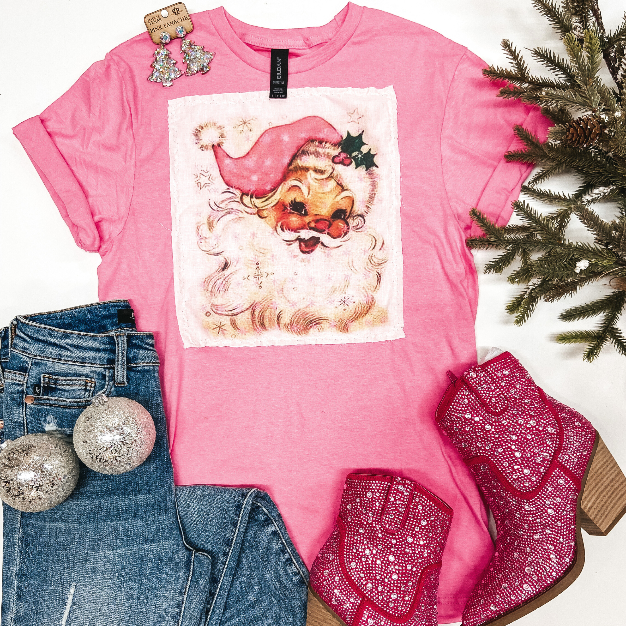 A pink short sleeve tee shirt with a white square patch sewn on the front with a vintage style image of Santa Clause. Pictured on a white background with garland, ornaments, silver earrings, pink booties, and a pair of folded jeans.