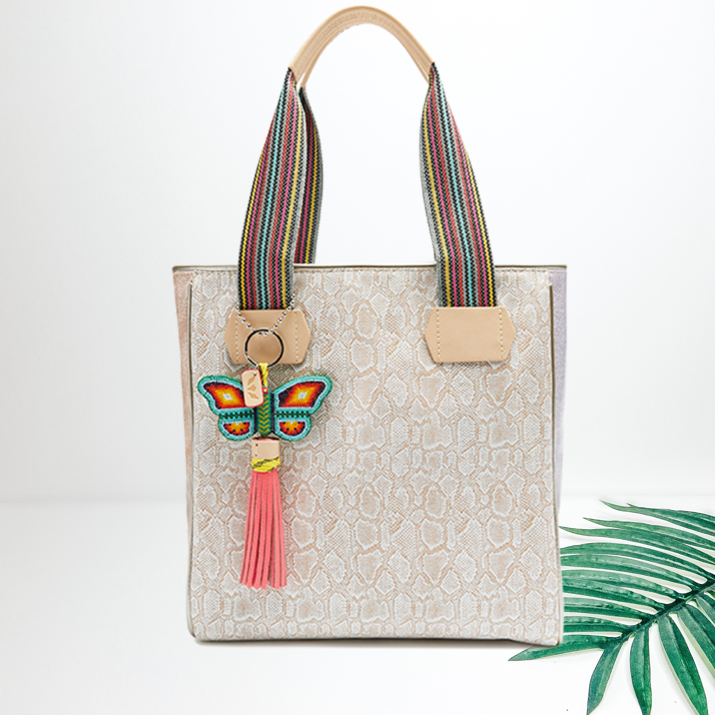 In the middle of the picture is a cream colored snake sin tote with a pink tassel butterfly charm. To the right of the tote is a green palm left on a white background.
