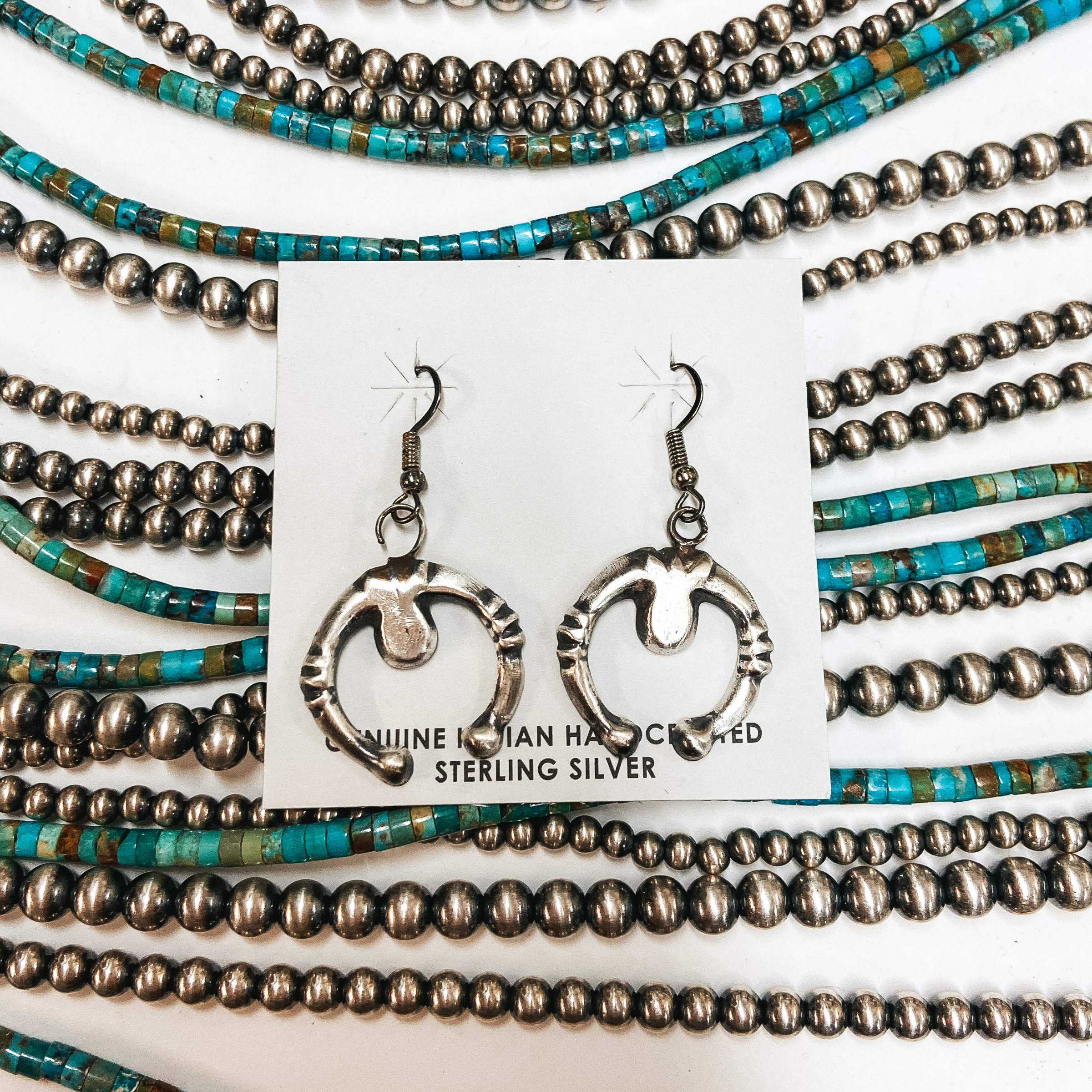 A pair of sterling silver naja shape earrings on a card displaying "Genuine Indian Handcrafted Sterling Silver." Pictured on white background with Navajo Pearls and turquoise beads.