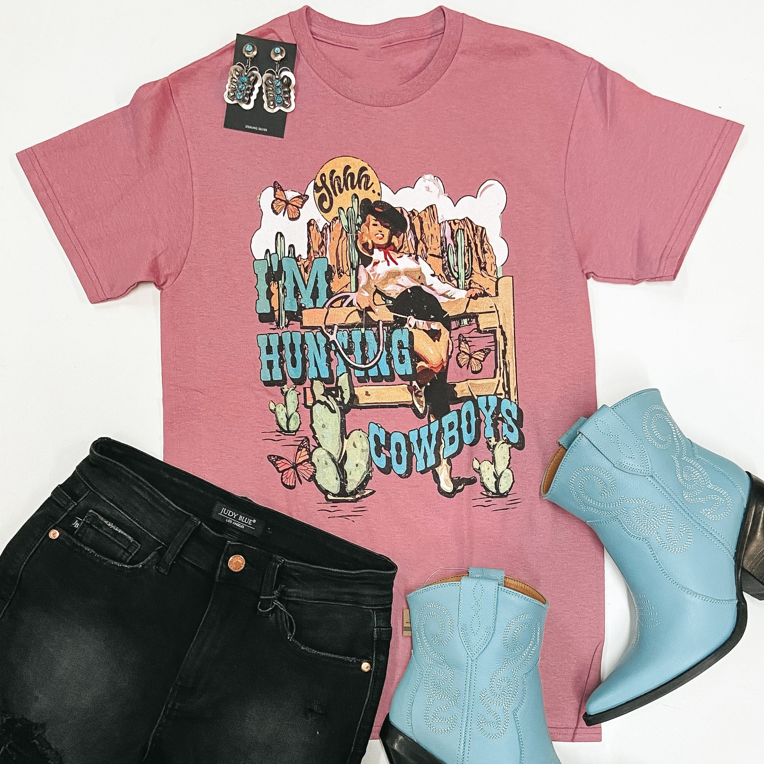 A mauve pink graphic tee is pictured on a white background. This short sleeve tee shirt has a graphic on the front center that is a pin-up cowgirl that says "Shhh.. I'm Hunting Cowboys." 