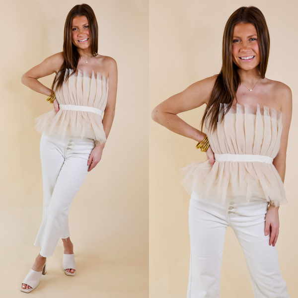 Model is wearing a strapless top made of tulle ruffles that is natural off-white. Model has this top paired with white jeans, ivory heels, and gold jewelry.