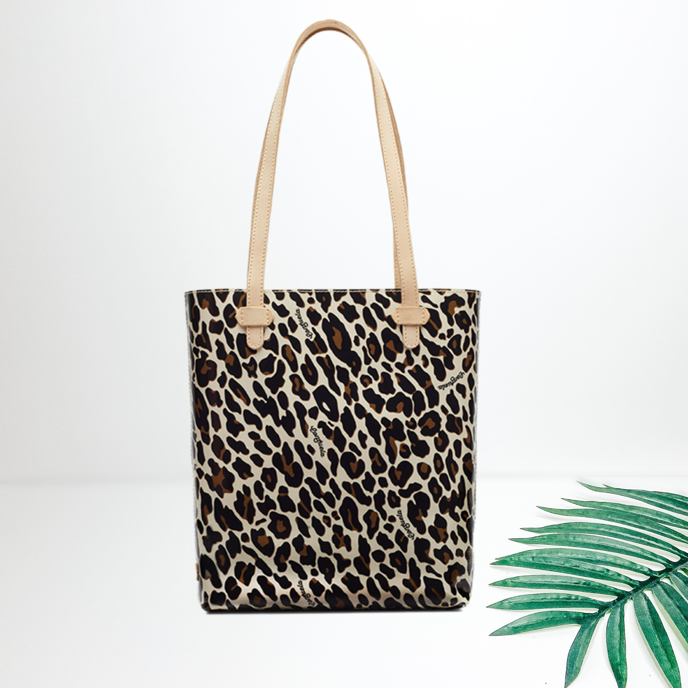 Centered in the picture is a tote bag in brown cheetah. To the right of the tote is a palm leaf, all on a white background.