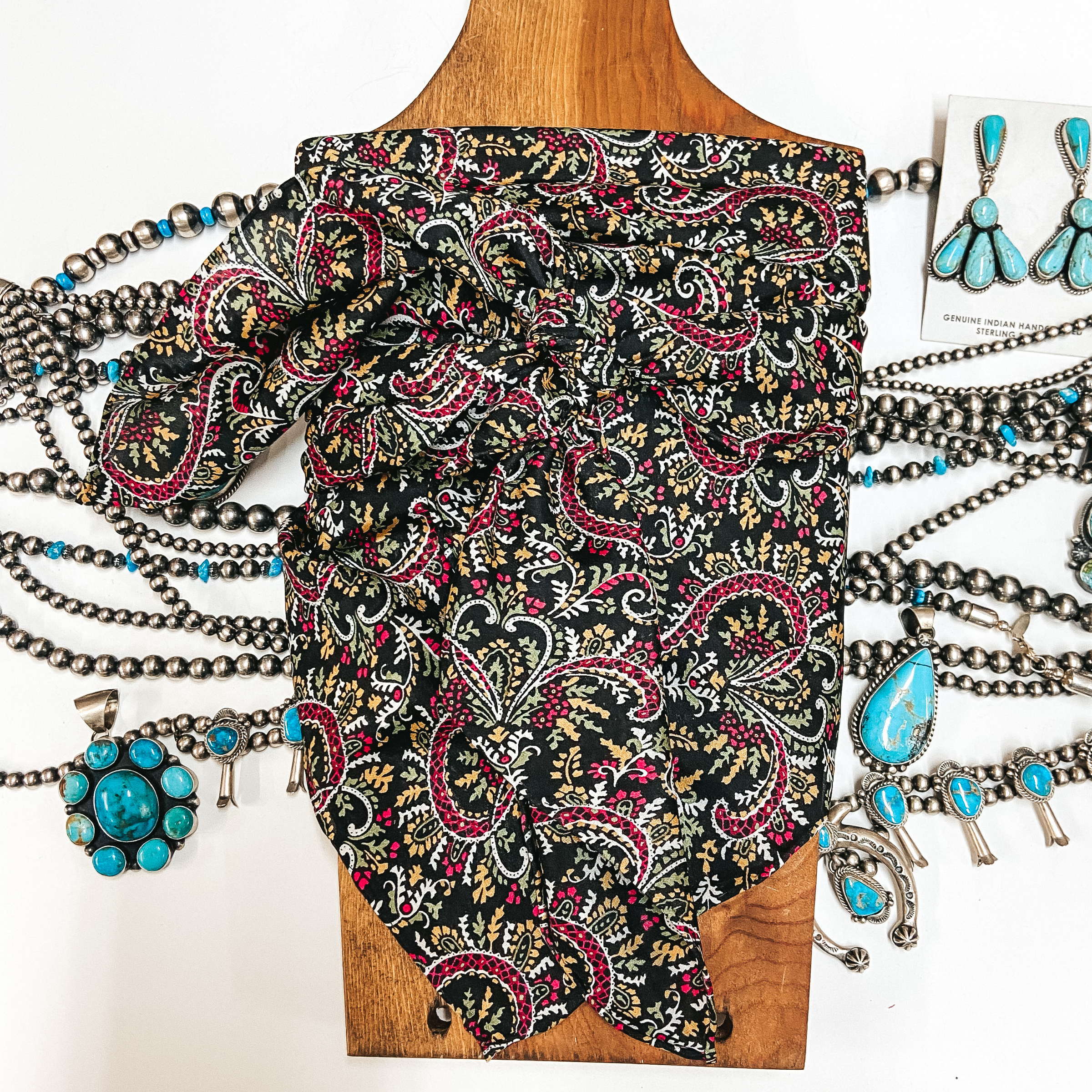 A black wild rag with multi colored paisley print tied around a wooden display. Pictured on white background with silver and turquoise jewelry.