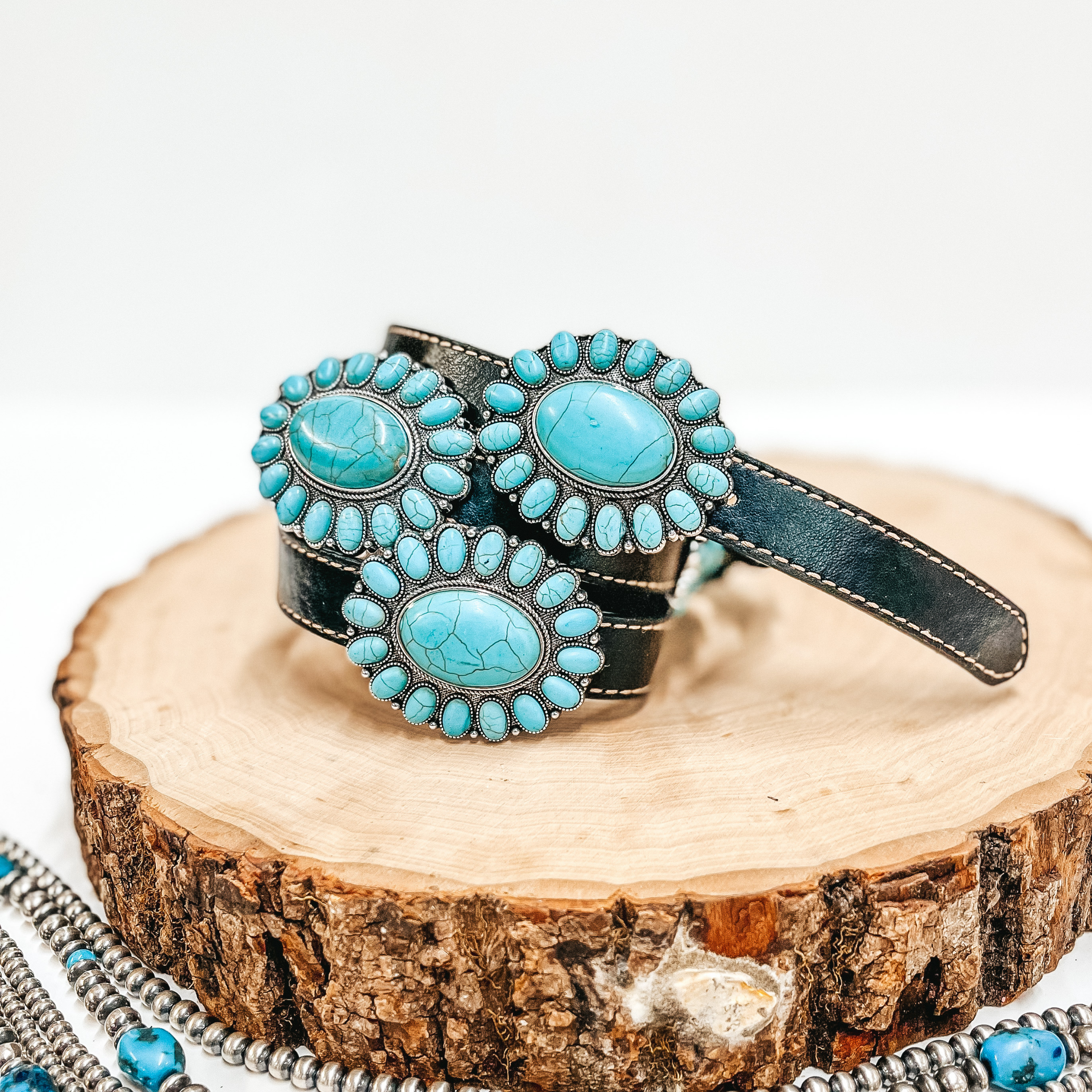 A black belt with faux turquoise cluster conchos pictured on wooden display with Navajo pearls on a white background.
