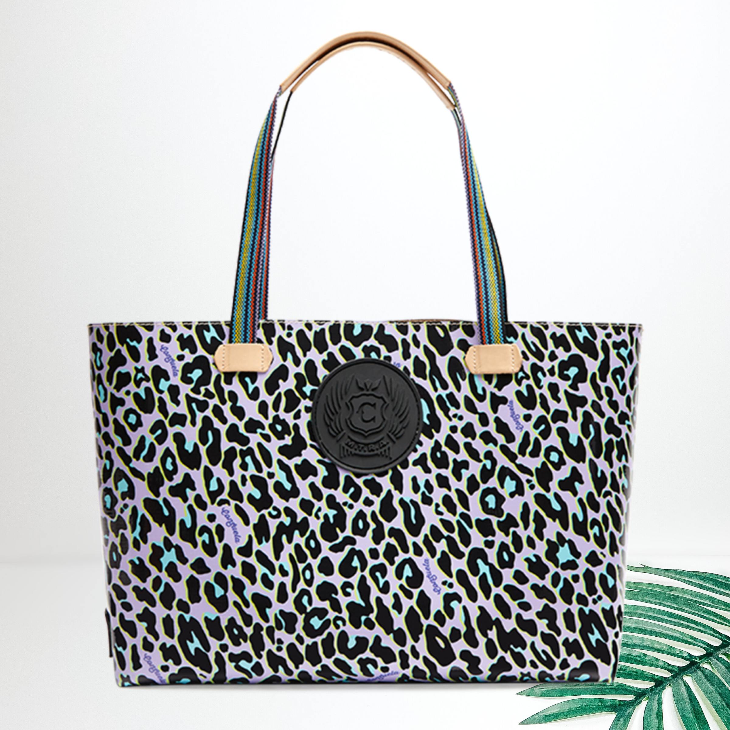 A large purple leopard print tote with a black branded patch. Pictured on white background with a palm leaf.