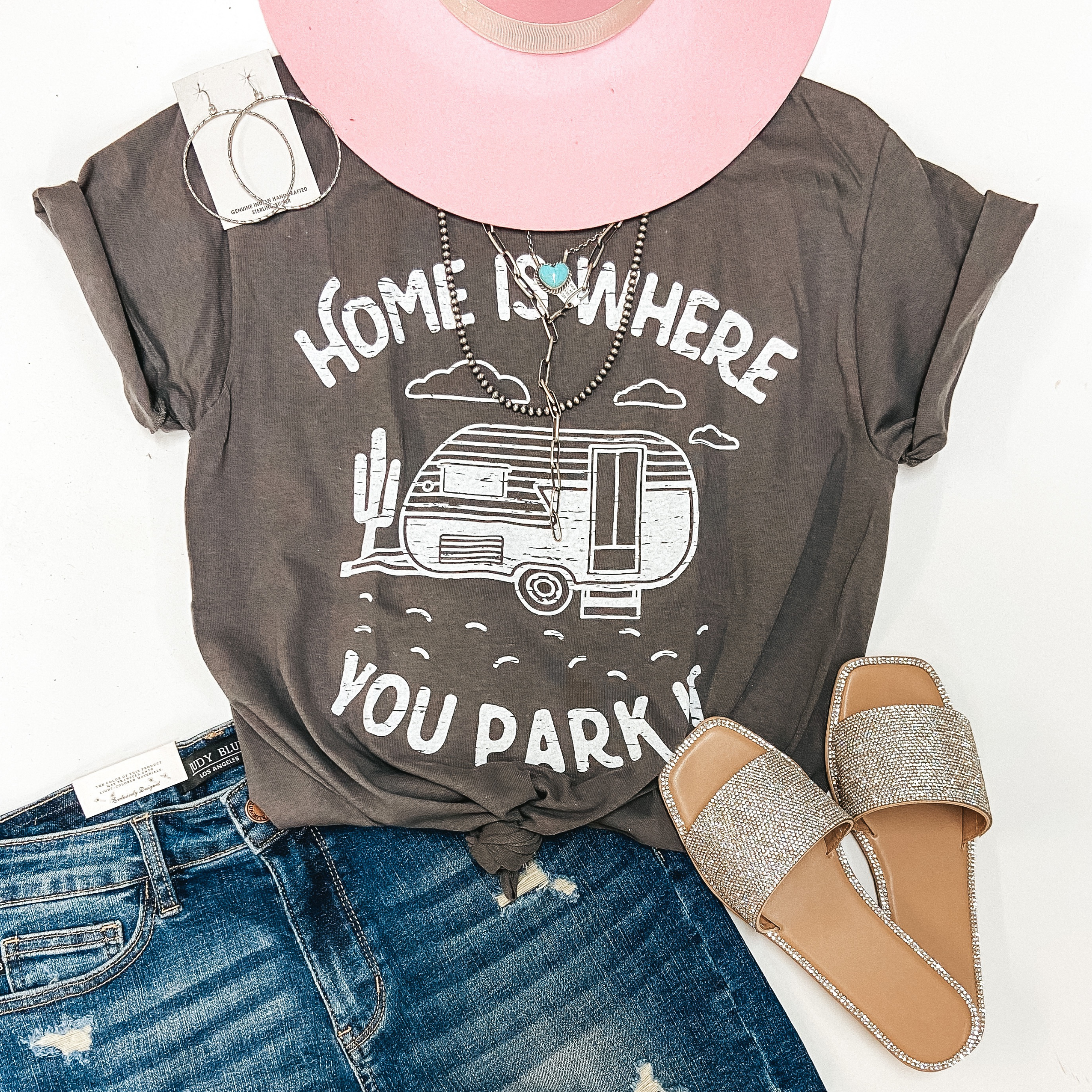A grey graphic tee that has the sleeves rolled and a knot on the front. The tee shirt has a camper and cactus graphic and says "Home Is Where You Park It." Tee shirt is pictured on white background with a pink hat, silver jewelry, denim shorts, and crystal sandals.