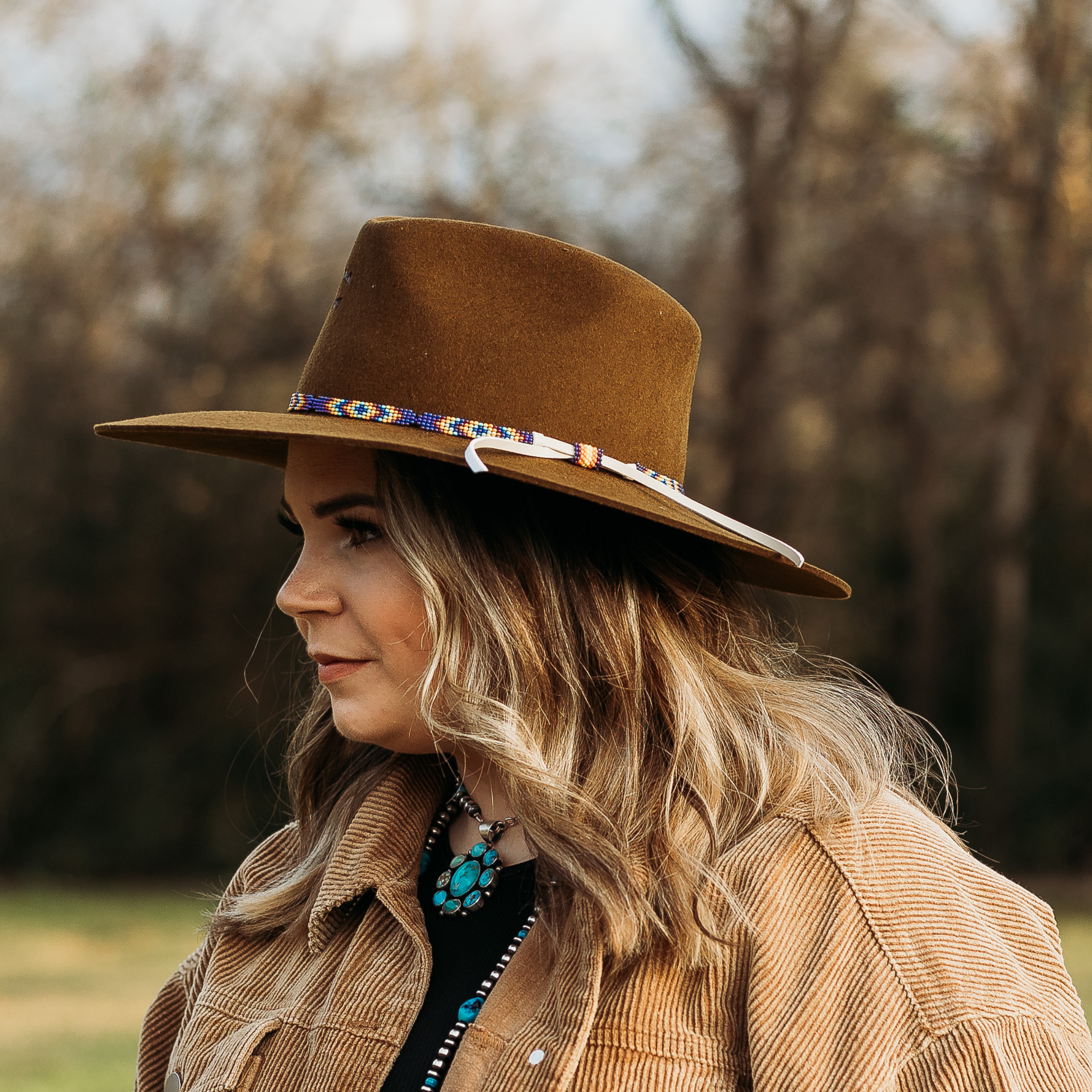 Tan felt hat with a Aztec beaded band. Model has it paired with a brown jacket and turquoise jewelry. Pictured on wooded background.