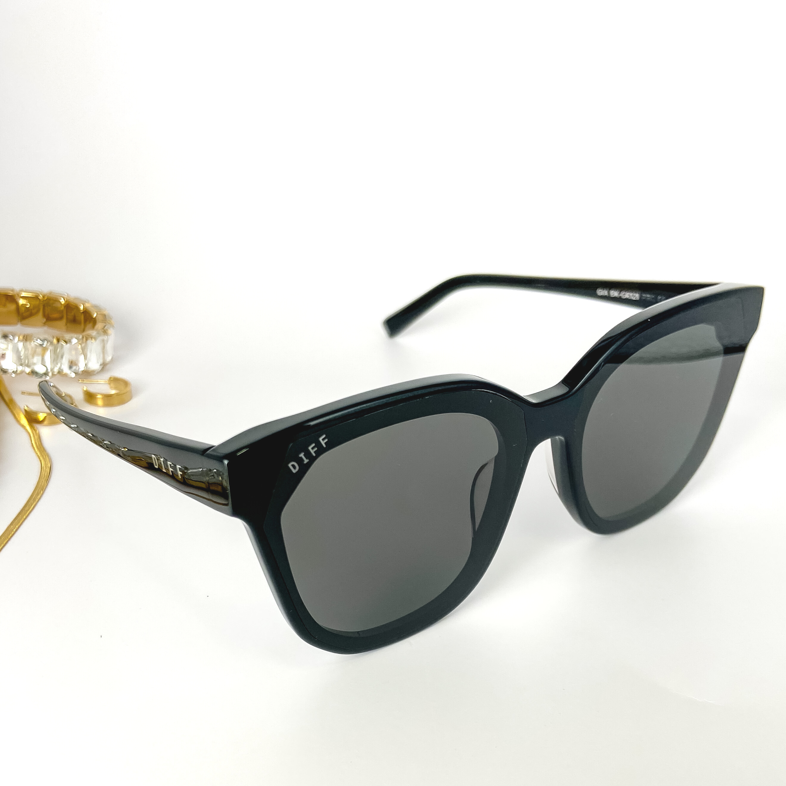 A pair of black cat-eye sunglasses with black lenses. These sunglasses are pictured on a white background with gold jewelry.