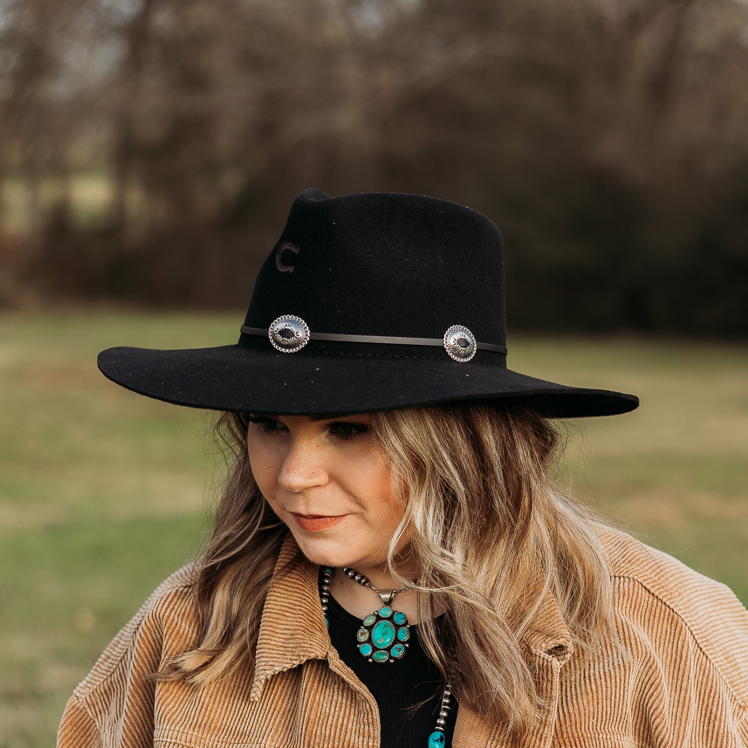 Black Felt Hat with Floppy Brim. Thin Black Leather Hat Band With Silver Conchos Along It. Model has it paired with a brown jacket and turquoise jewelry. Pictured on wooded background.