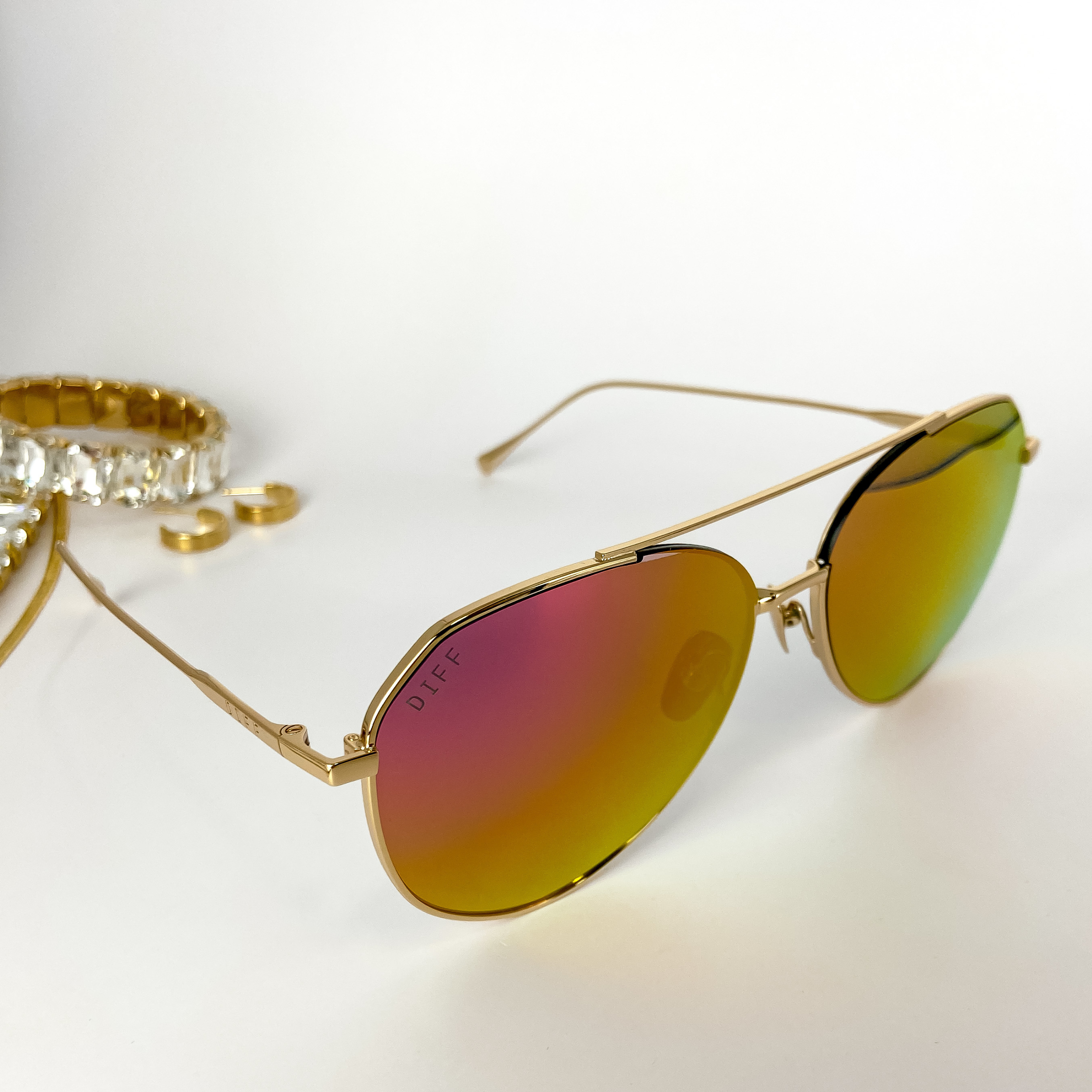 A pair of gold-tone aviator sunglasses with pink and gold mirror lenses. These sunglasses are pictured on a white background with gold jewelry.