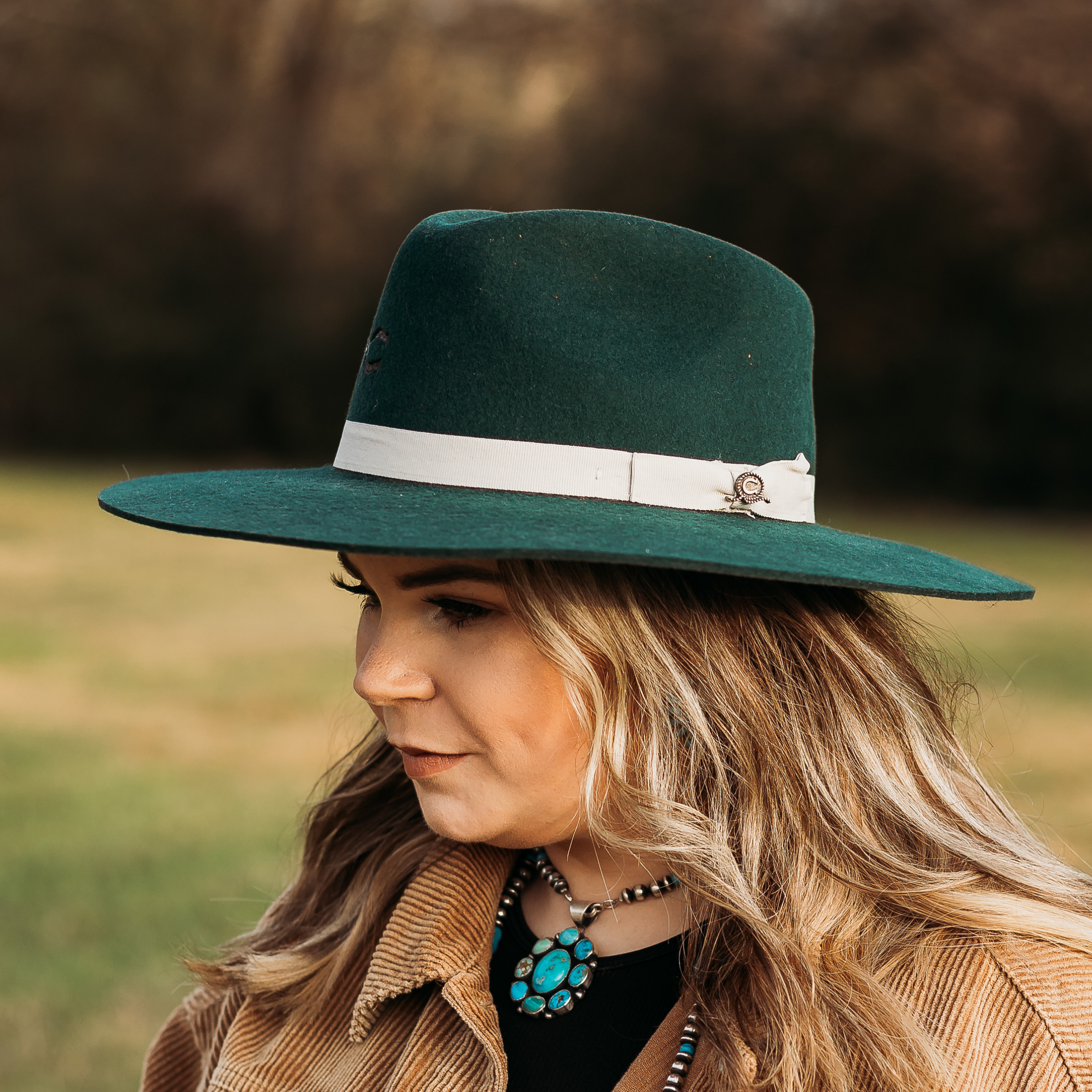 Forest Green Felt hat with White Hat Band and Charlie 1 Horse Pin. Model has it paired with a brown jacket and turquoise jewelry. Pictured on wooded background