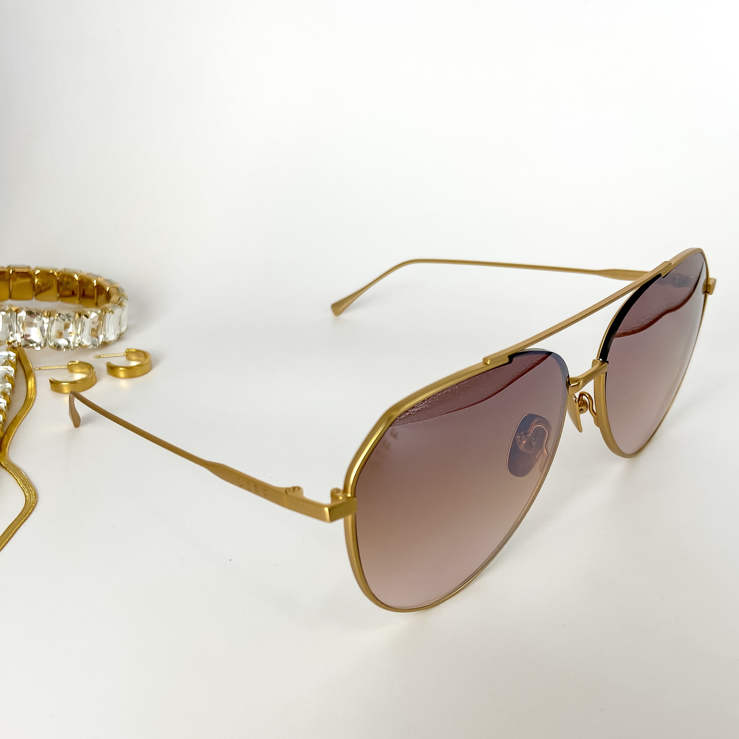 A pair of gold tone aviator sunglasses with taupe mirror lenses. These sunglasses are pictured on a white background with gold jewelry.