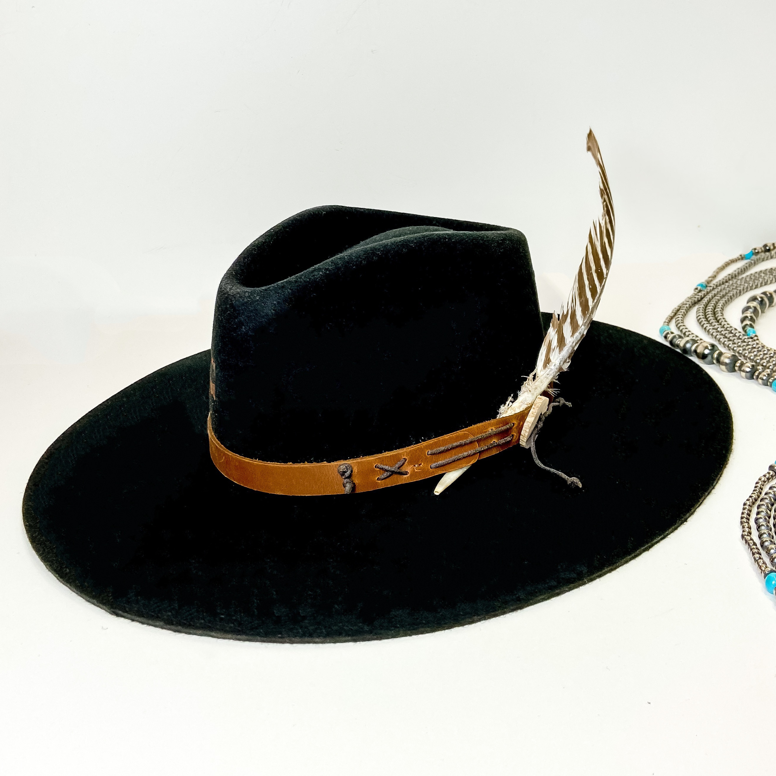 A black felt hat with a leather band and threaded detailing with a feather. This hat is pictured on a white background with genuine Navajo pearls.