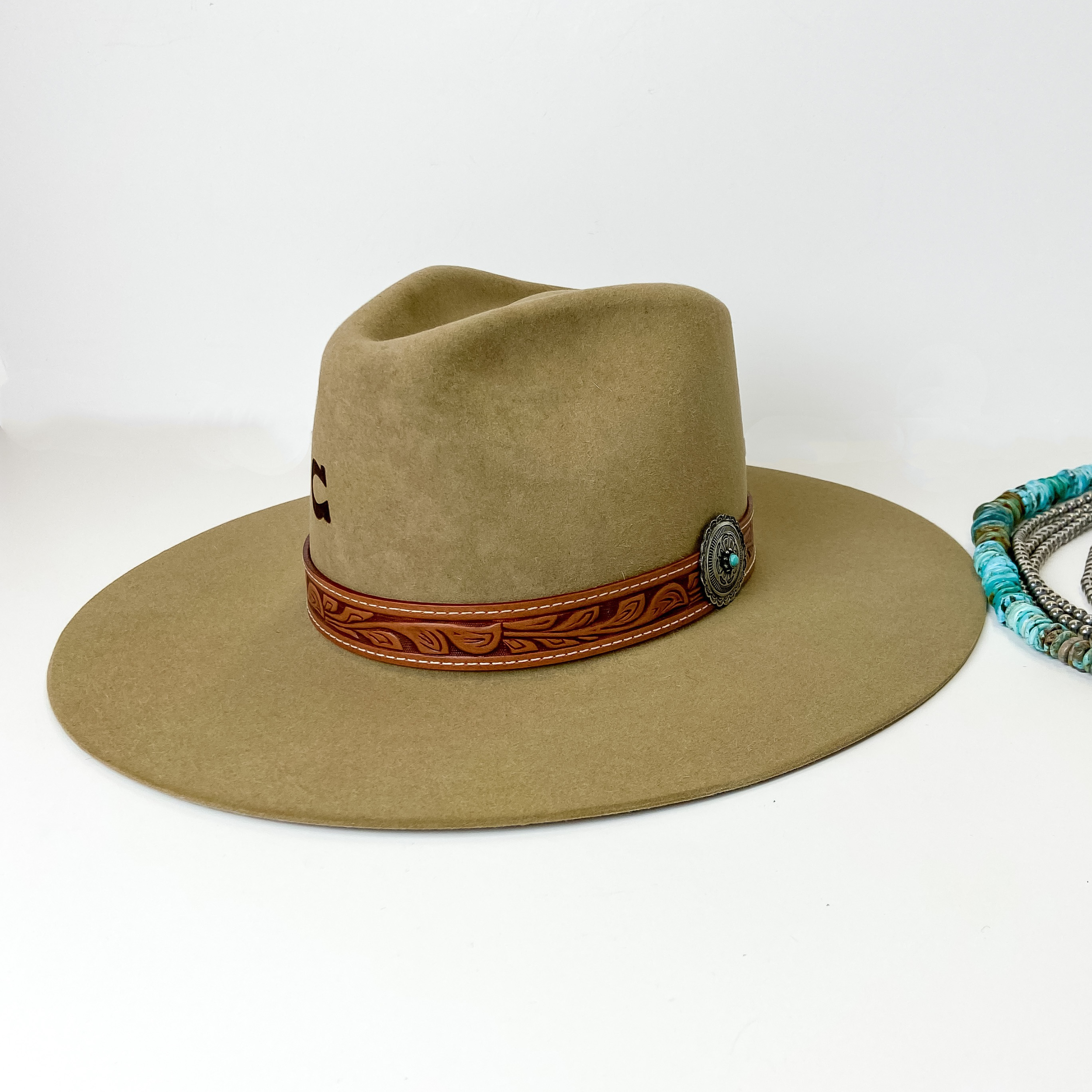 A light brown felt hat with a pinched front, flat brim, and a tooled leather band with a silver tone concho and turquoise stone. This hat is pictured on a white background with genuine Navajo jewelry.