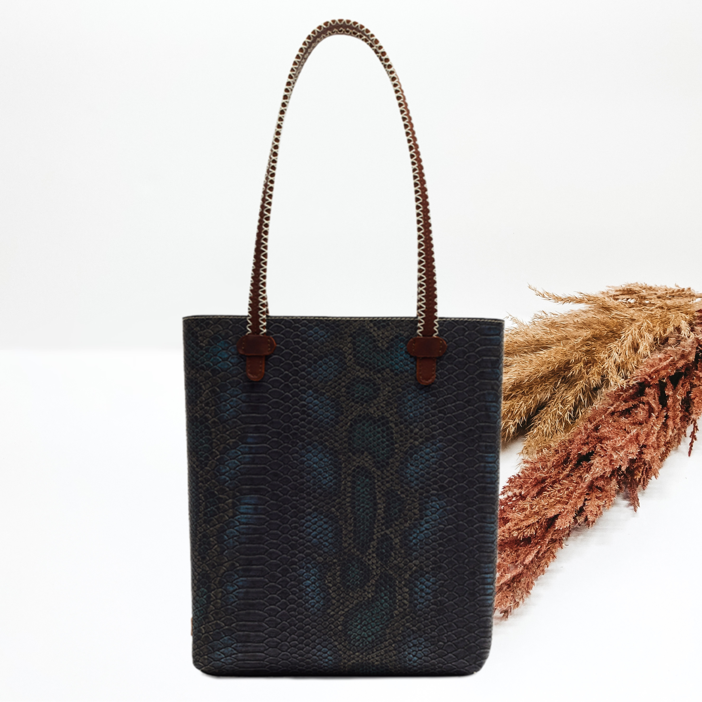Rectangle tote bag with a dark snake skin print and leather stitched straps. This tote is pictured on a white background with pompous grass behind the tote. 