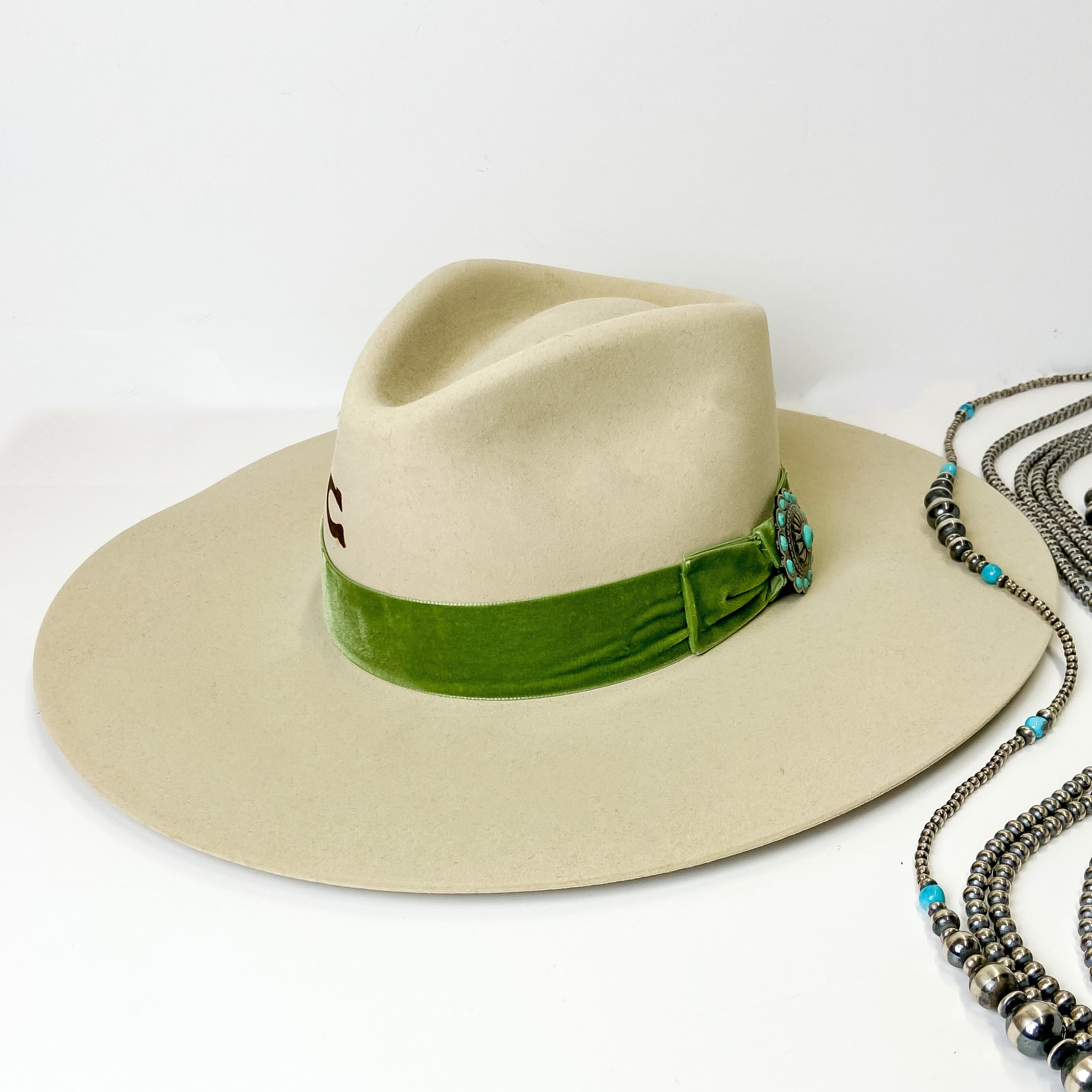 An ivory felt hat with a green velvet band with a silver tone concho with turquoise stones. This hat is pictured on a white background with genuine Navajo jewelry.