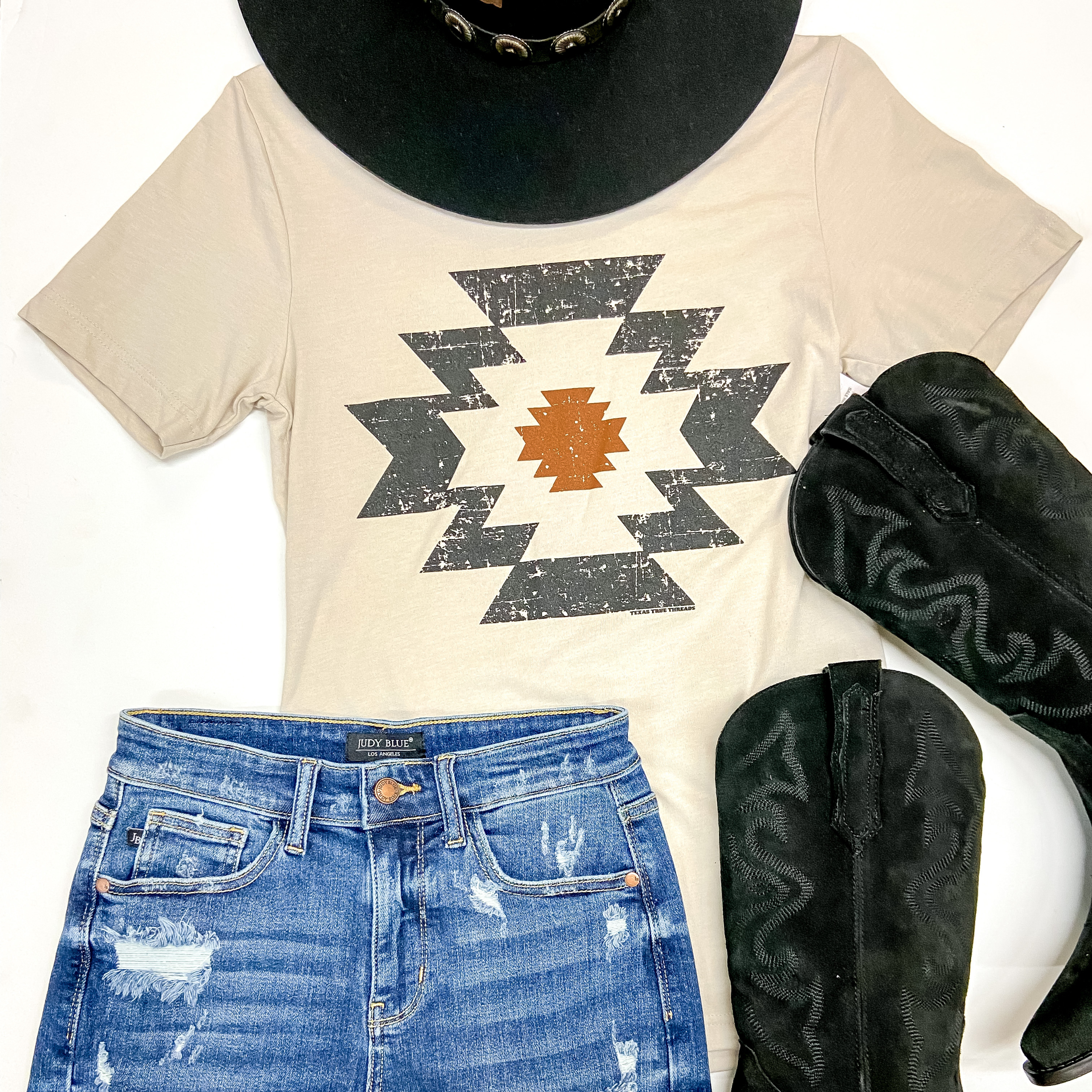 A beige short sleeve graphic tee with a black and orange tribal symbol graphic. This tee shirt is pictured on a white background with black boots, a black hat, and denim shorts.