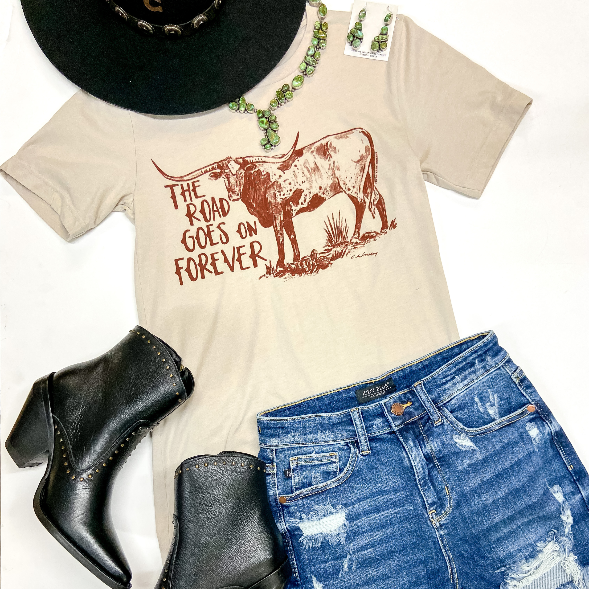 A beige short sleeve graphic tee with a long horn and the phrase "The Road Goes on Forever." This tee is pictured on a white background with denim shorts, black booties, and genuine turquoise jewelry.